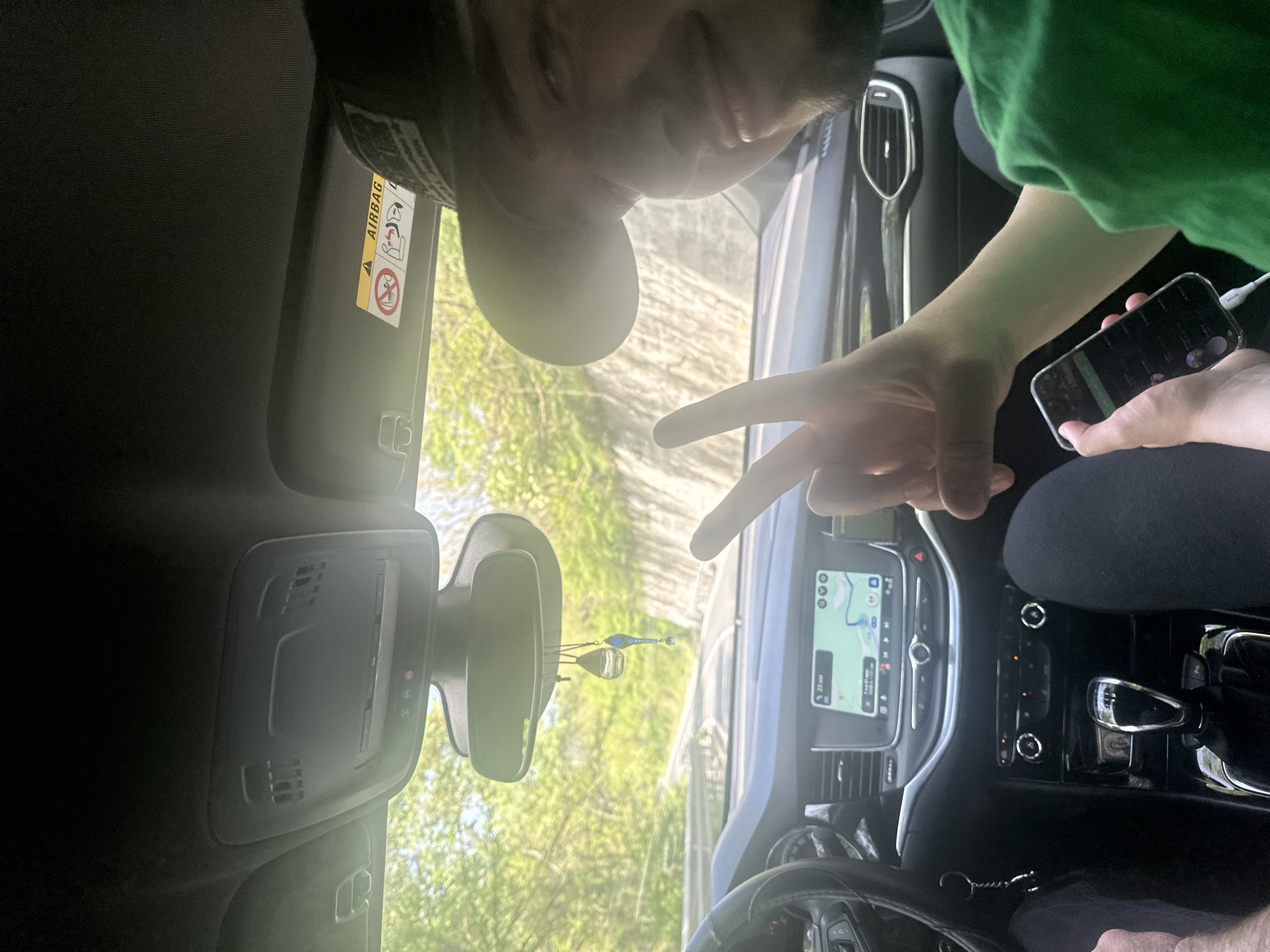 The image depicts the interior of a car. A person wearing a black cap and another person with a green shirt are visible. One of them is holding a smartphone. The car's dashboard features a navigation screen displaying a map. A pair of decorative items are hanging from the rearview mirror. The location is identified as Bilka, Bulgaria.