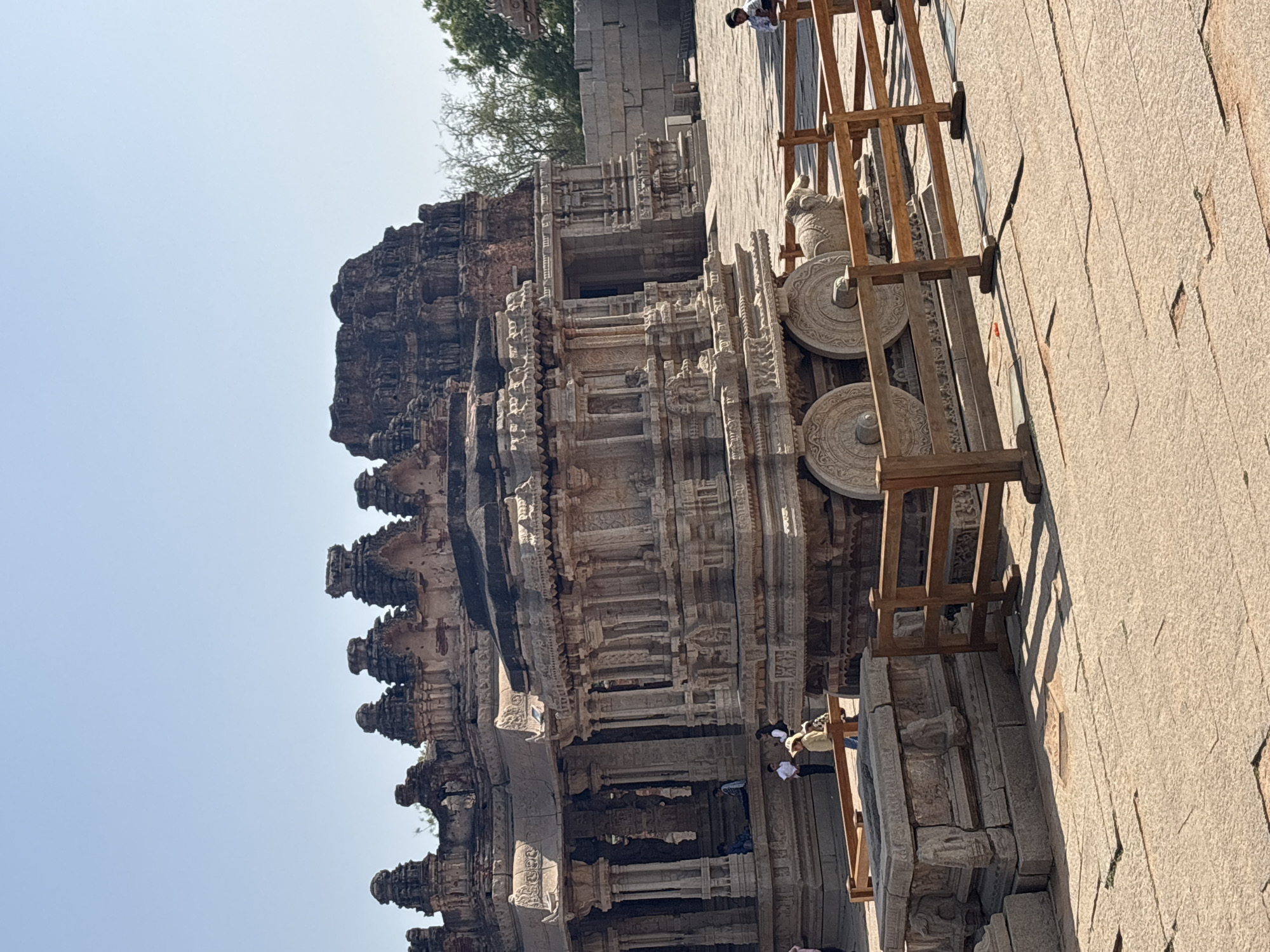 The image shows the Stone Chariot at the Vittala Temple complex in Hampi, India. The chariot, a monolithic structure, is made of stone and features intricately carved wheels. A wooden fence surrounds the chariot, likely to protect it from damage. Two people are visible, both wearing white shirts, one standing near the base of the chariot and the other standing on the stone floor. The stone floor is light colored and has a smooth, worn texture. The sky is clear and light blue. The architecture is ancient and characterized by detailed carvings. The picture is taken during the daytime under bright sunlight, casting shadows on the structures.