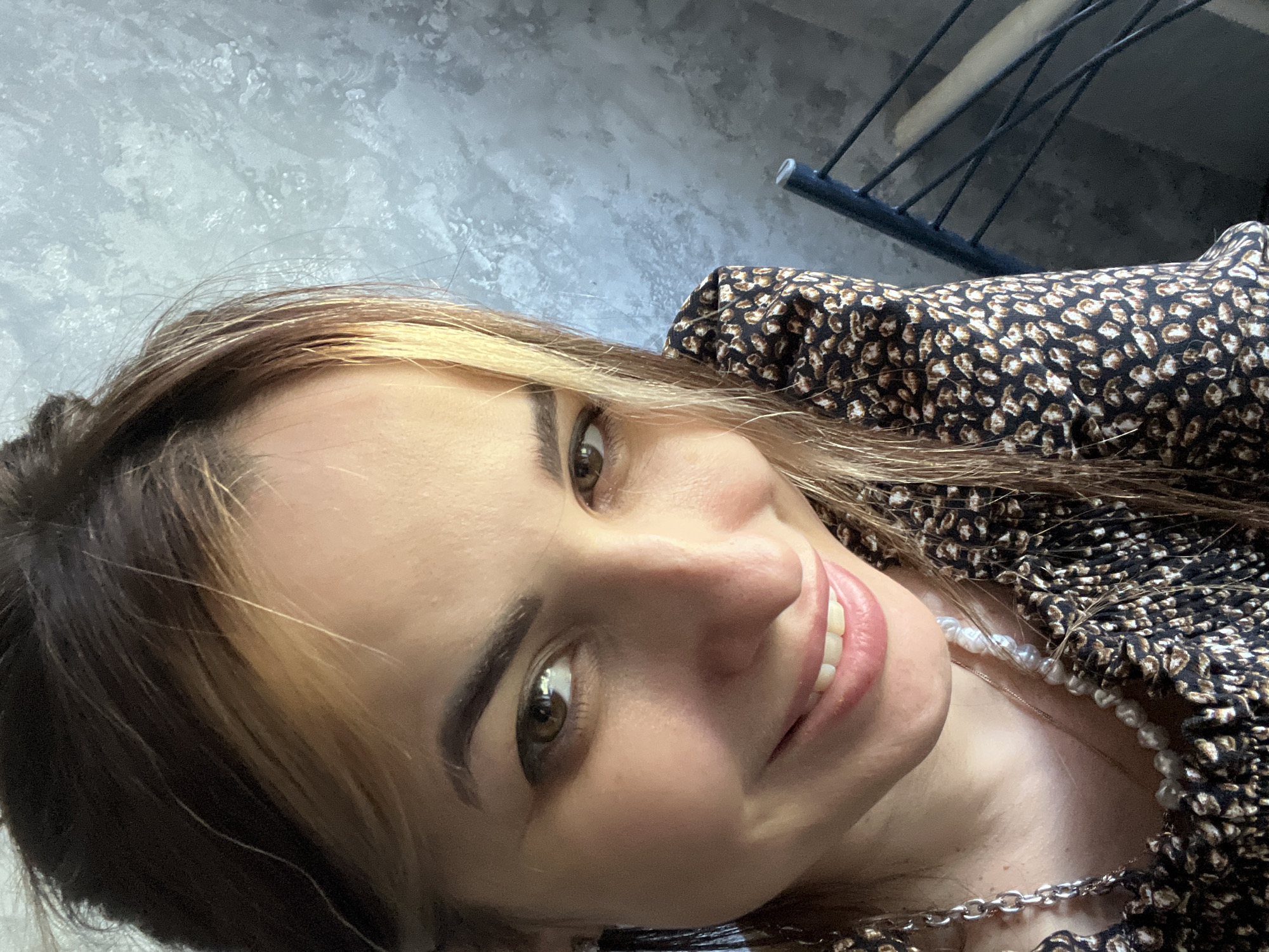 A close-up, slightly rotated headshot of a smiling woman is presented. The woman has brown hair with blonde highlights framing her face. Her eyes are brown and she is wearing light pink lipstick. A small portion of a black and brown patterned top is visible. A strand of pearls and a silver chain necklace are also visible. The background consists of a textured gray wall and a portion of a black metal structure. The location is unknown.
