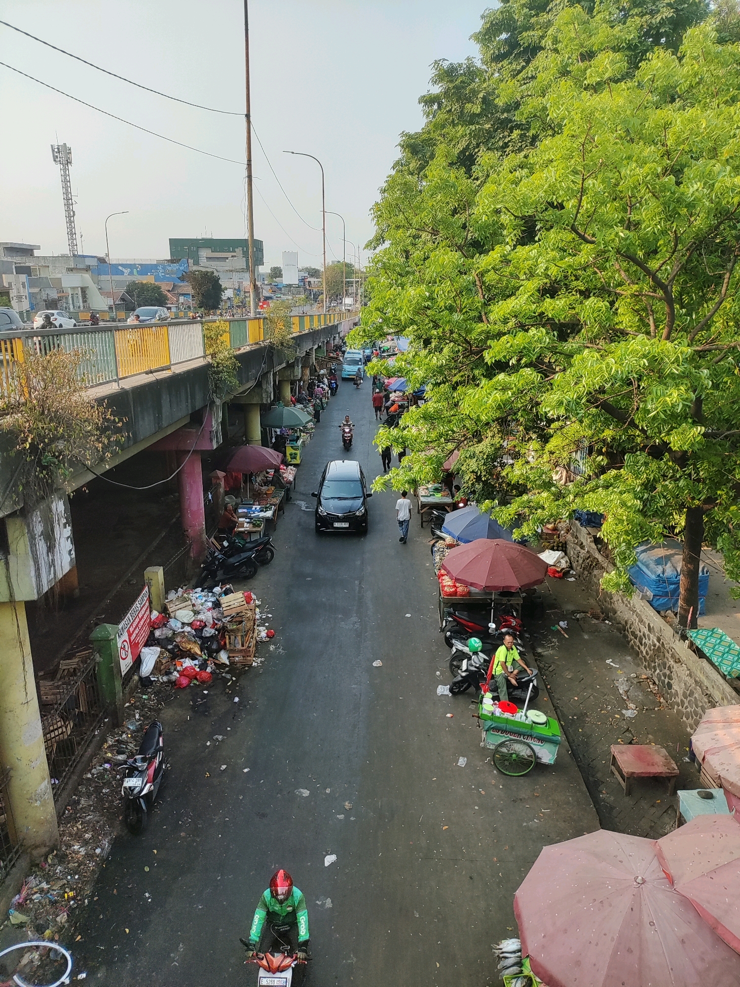 The image depicts a street scene in East Jakarta, Indonesia, likely a market area under an overpass. The road is wet and littered with debris. On the left side of the road, under the overpass, is a large pile of garbage bags and wooden crates, some overflowing. A green sign with red lettering is partially visible in front of the garbage pile. A motorcycle is parked near the garbage.
On the right side of the road, there are several market stalls with vendors and customers. Red and blue umbrellas are open over some of the stalls. A vendor is sitting on a motorcycle at one of the stalls. There's a food cart with an umbrella. Several motorcycles are parked along the roadside. In the background, a black car is driving down the road, and further up, more vehicles are visible, including a blue bus. A person on a motorcycle, wearing a green jacket and helmet, is riding away from the camera. Trees with lush green foliage line the right side of the street, extending over the market stalls. The overpass structure is prominent, with concrete pillars supporting it. In the distance, buildings and a telecommunications tower can be seen. The sky is bright and clear, suggesting daylight.