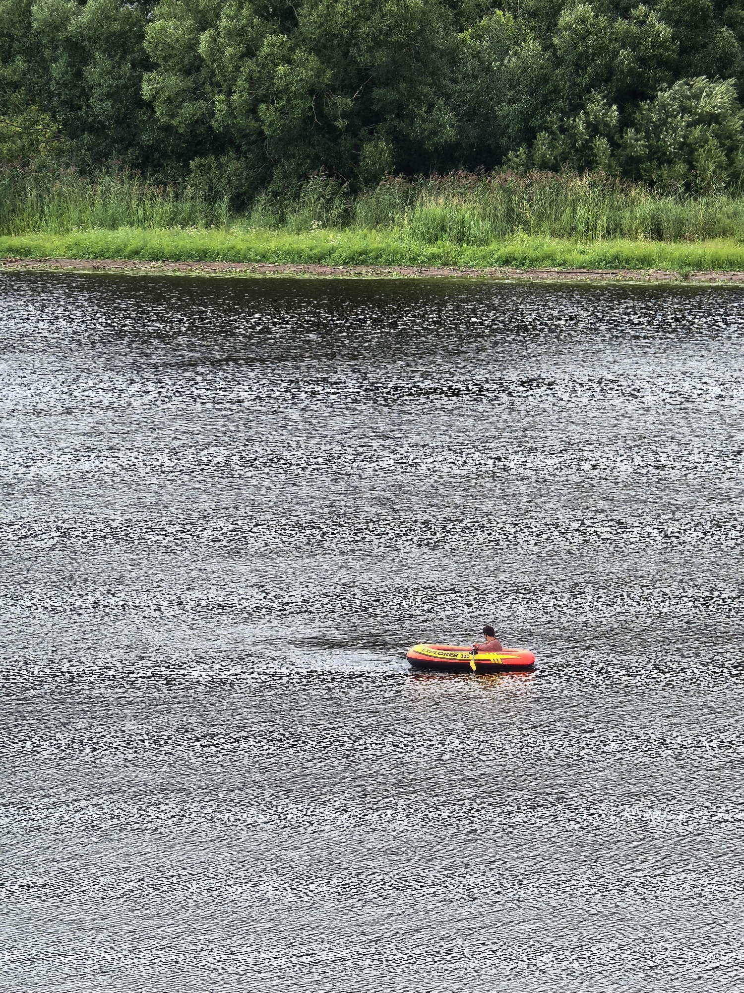 A person is in a yellow and red inflatable boat on a dark, rippling body of water. The person is wearing a reddish-brown shirt and is holding a yellow oar. The boat has the words "EXPLORER 300" written on its side. In the background, a lush green treeline runs along the bank of the water. The location is identified as Staraya Ladoga, Russia.