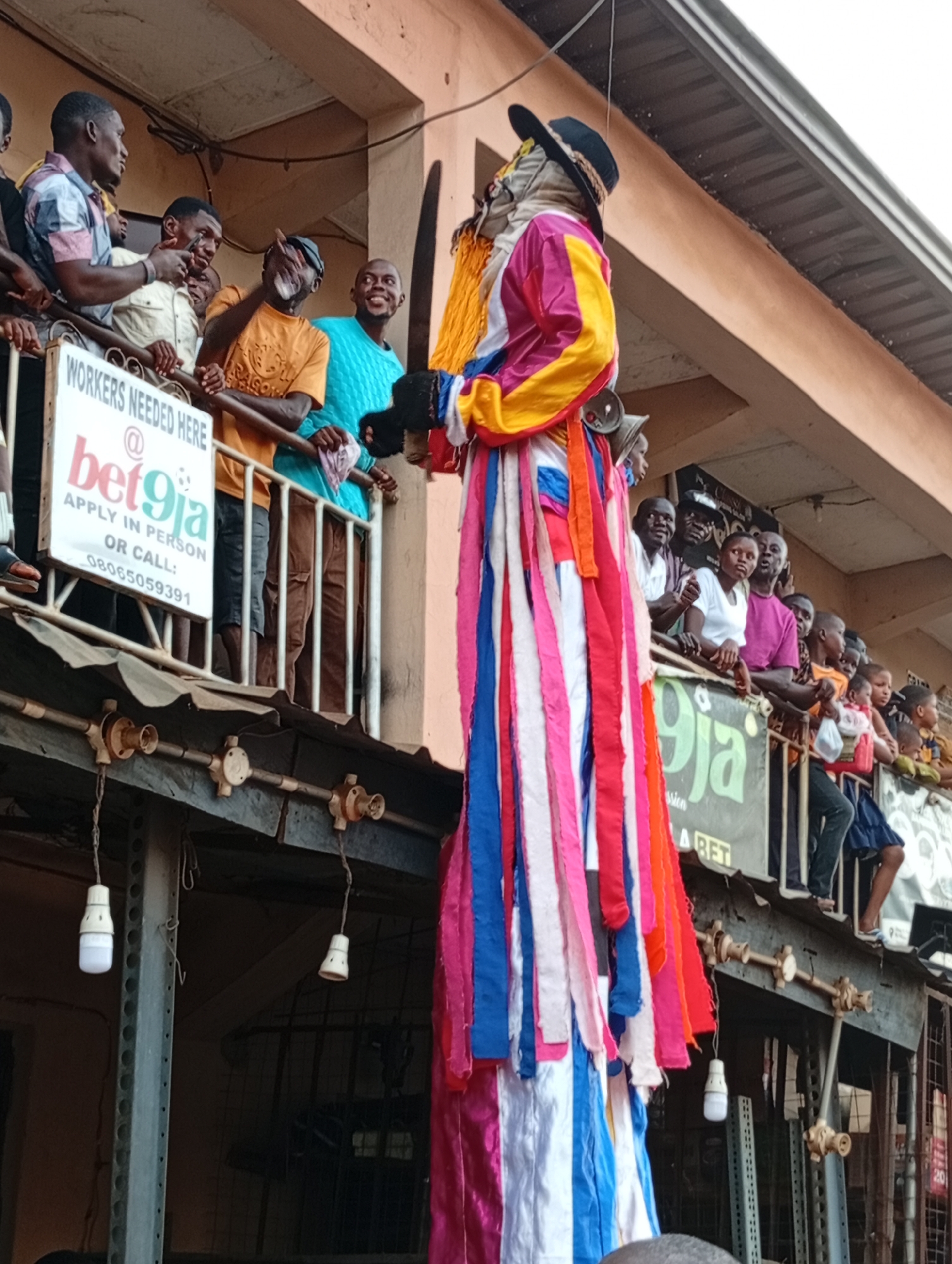 Here is a detailed description of the image:
**Scene:** An outdoor scene in a city of Unknown, Unknown shows a crowded balcony or veranda. A tall figure in elaborate colorful garb stands out, attracting the attention of the people on the balcony.
**People:** The balcony is populated with several people, ranging from adults to children, observing the figure. They appear to be a mix of men and women. A few individuals appear to be smiling or gesturing towards the figure.
**Subjects/Objects:**
* The tall figure is adorned with a costume of many colors, including red, yellow, blue, white, and pink ribbons or streamers. It also has a black hat.
* Two bells can be seen around the figure's neck.
* A sign for "bet9ja" is affixed to the balcony railing, indicating it could be a commercial space.
* Visible beneath the balcony is a corrugated metal roof, with hanging light bulbs.
**Actions/Interactions:**
* The people on the balcony are mainly observing the figure below.
* Several of the people are engaging with each other, talking or smiling in the direction of the figure.
**Environment:**
* The scene appears to be in an urban area, likely a market or commercial street, given the balconies.
* The weather is somewhat overcast.
* The location is probably in Africa, going by the race of the people present.
The overall impression is of a local celebration or event.