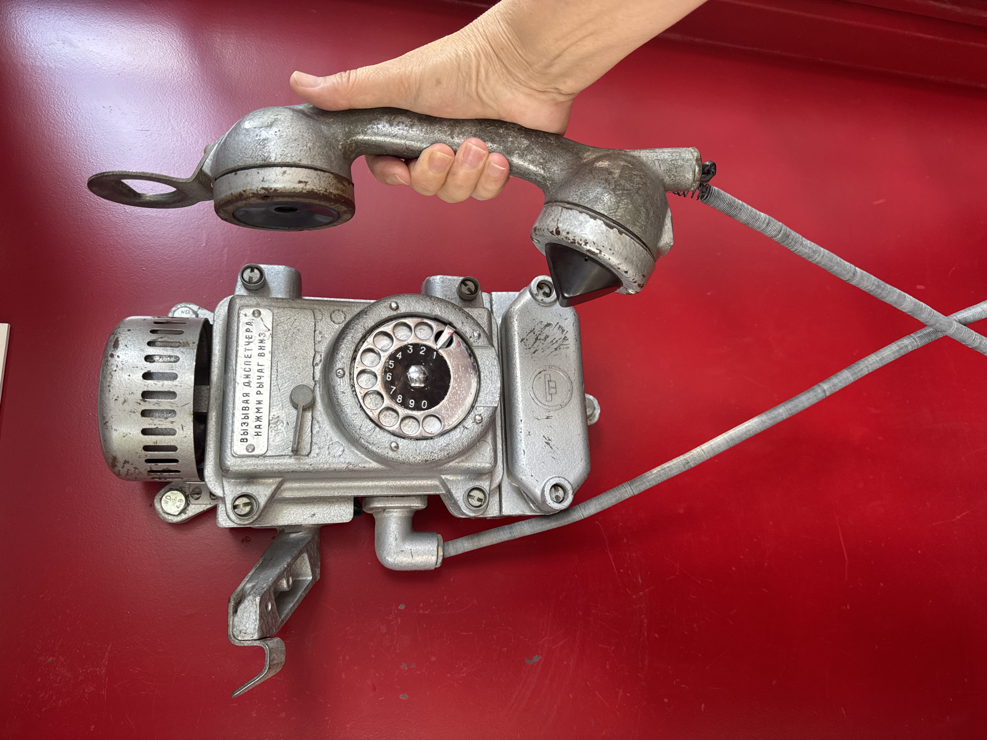 A vintage, silver-colored rotary dial telephone, likely of Soviet or Russian origin given the Cyrillic inscription, is mounted on a bright red surface, possibly a wall or door. The inscription on the phone reads "ВЫЗЫВАЯ ДИСПЕТЧЕРА НАЖМИ РЫЧАГ ВНИЗ", which translates to "TO CALL THE DISPATCHER PRESS THE LEVER DOWN". A hand with visible fingers is holding the telephone receiver, with the receiver's earpiece end positioned near the phone's base. The telephone exhibits signs of age, with some scuffing and wear visible on its metal casing and the rotary dial. The red surface also shows minor scratches and dust marks. The scene appears to be an outdoor or semi-outdoor location, suggested by the texture and slight imperfections of the red background. The specific location in Kokshetau, Kazakhstan, is not discernible from the visual information present in the image.
