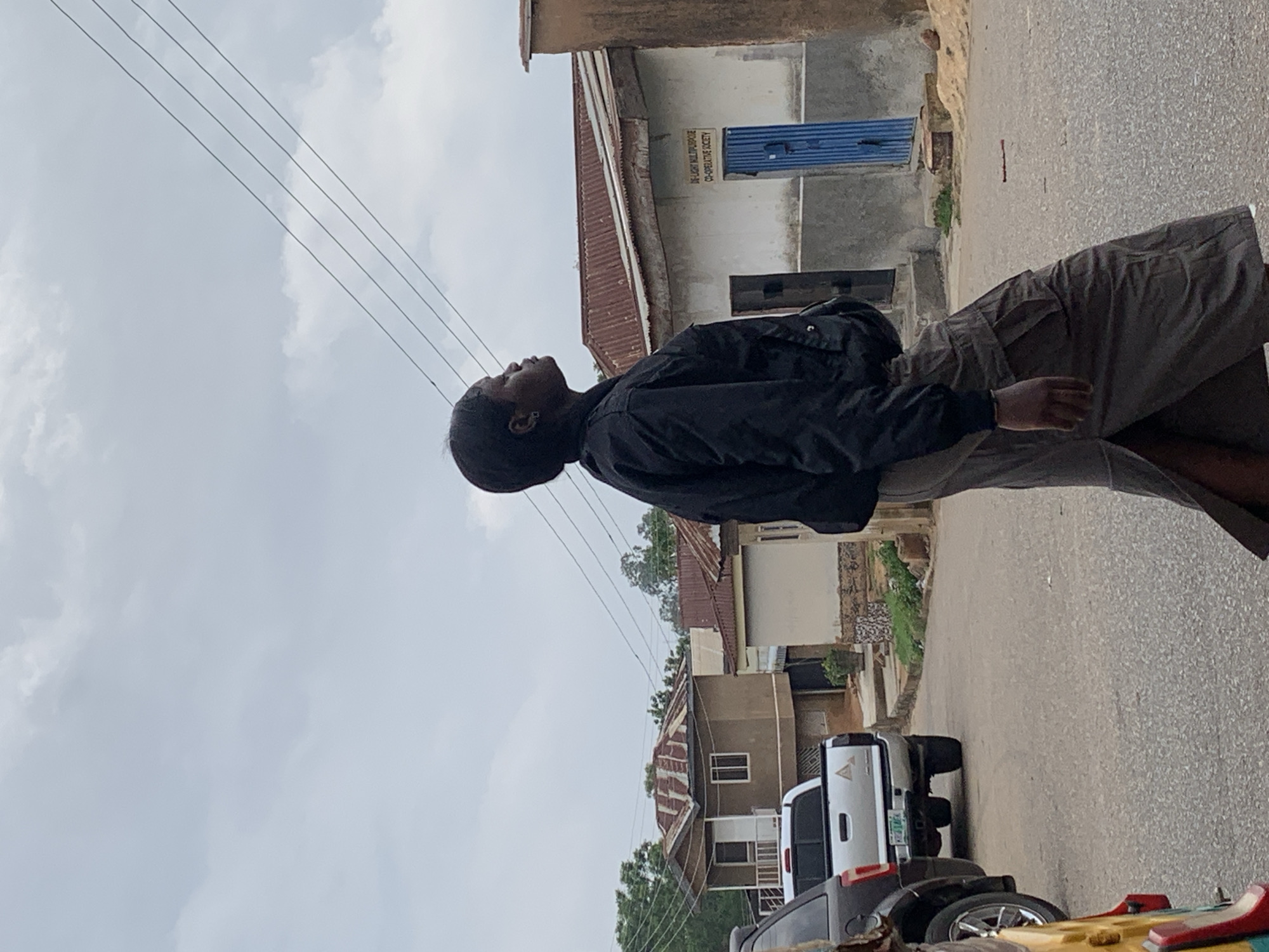 A person wearing a black jacket and grey trousers stands on a paved road in Jos, Nigeria, looking up towards the sky. Utility lines run overhead. In the background, residential buildings of varying heights and colors are visible, with the rooftops of some made of corrugated metal. A white pickup truck and other vehicles are parked along the roadside. A sign with blue accents is affixed to one of the buildings. The sky is overcast.