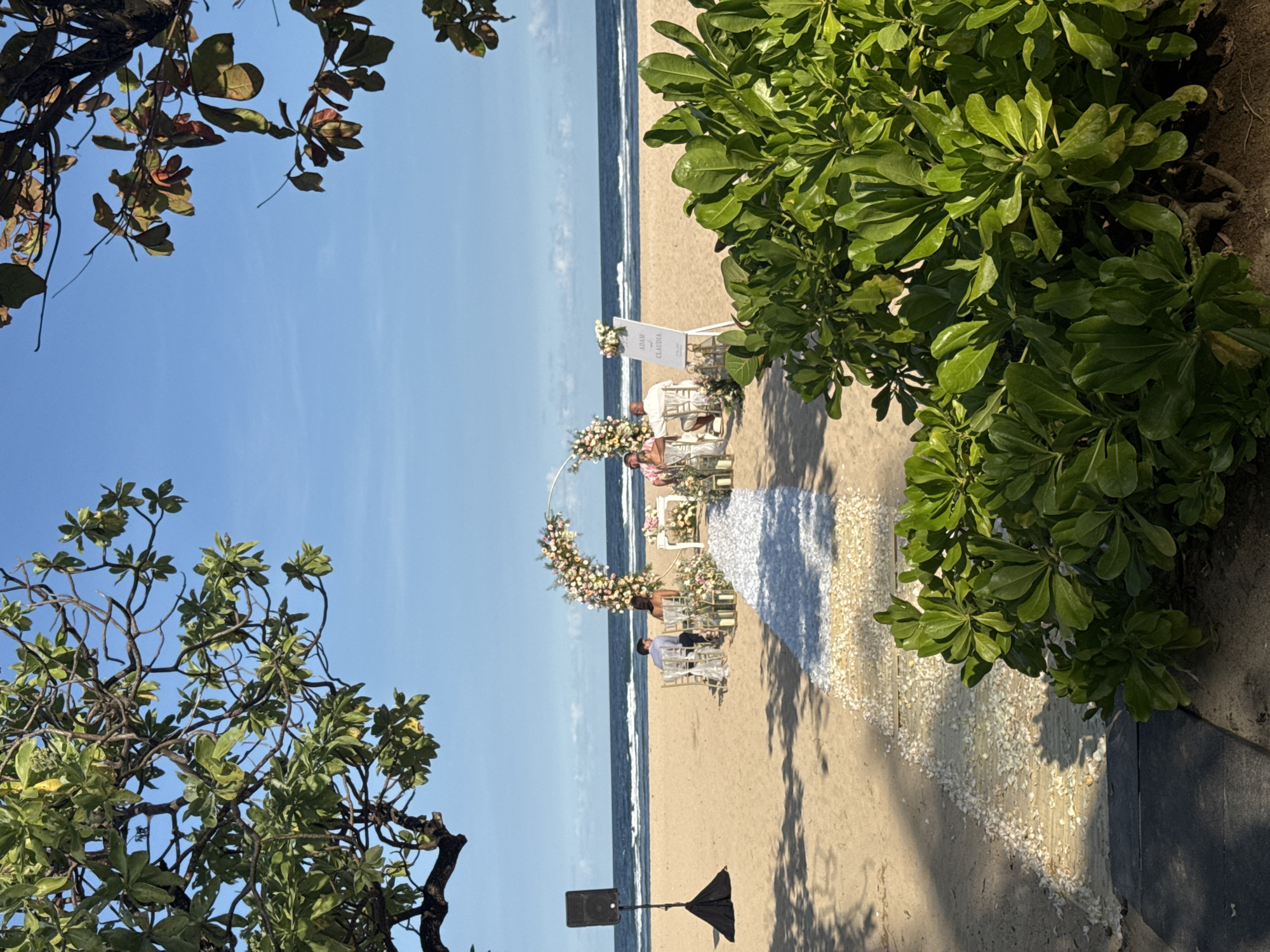 The image captures an outdoor wedding ceremony on a beach in Kuta Selatan, Indonesia. A couple stands under a floral arch facing the ocean, with white chairs positioned on either side of the aisle. The aisle is decorated with white petals, leading to the arch. A sign with the names "Adam & Claudia" stands near the couple. The scene is framed by trees on both sides, with a clear blue sky above. A black speaker is visible near the arch.
