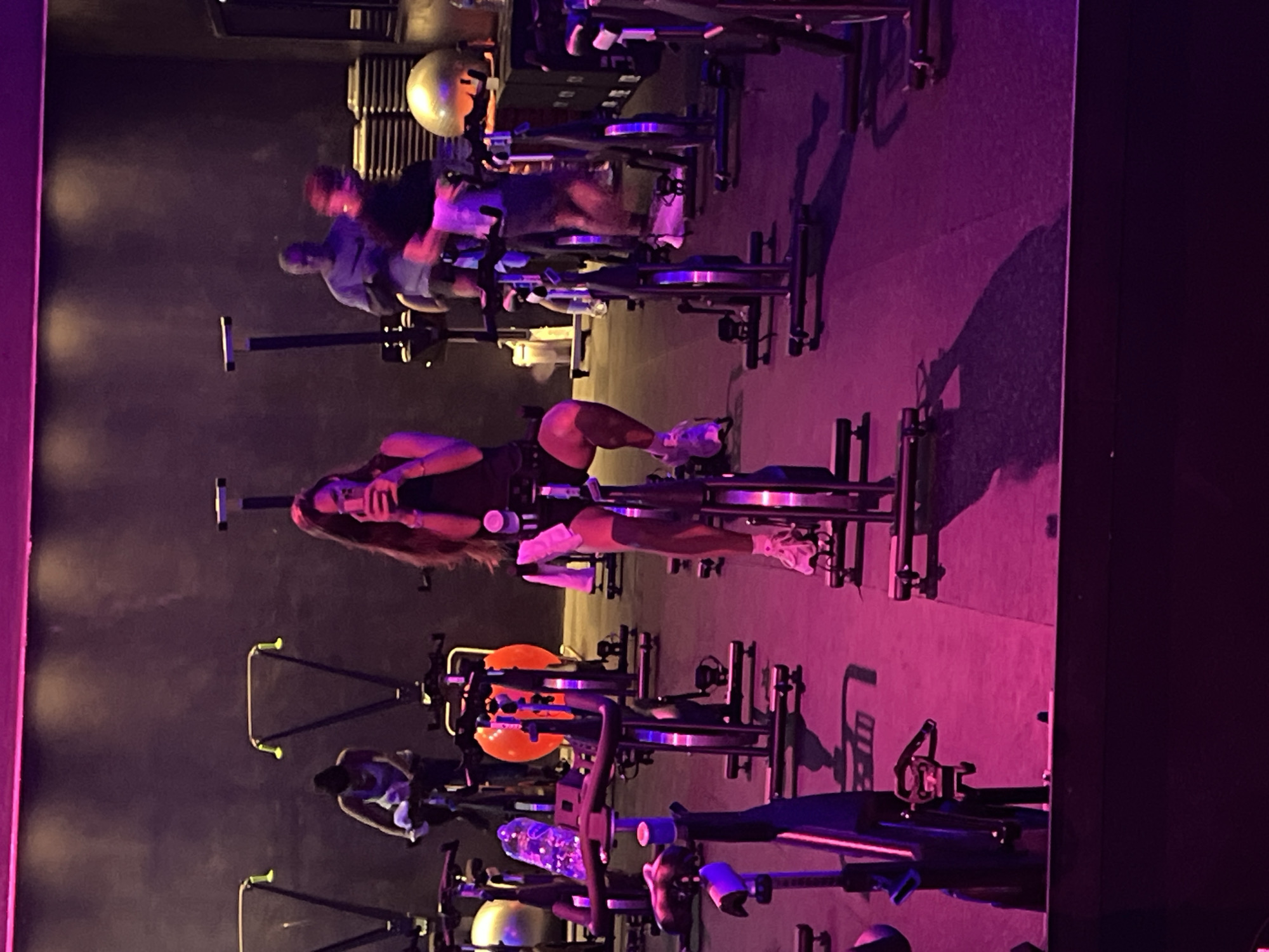 The image depicts an indoor cycling class in progress. Several stationary bikes are arranged in rows, occupied by individuals engaged in a workout. The lighting is predominantly purple and low, creating a fitness studio ambiance. One person is in the middle looking at a phone. Exercise equipment, including stability balls, is visible in the background. The location is indeterminate, and the image does not provide sufficient detail to ascertain a specific city.