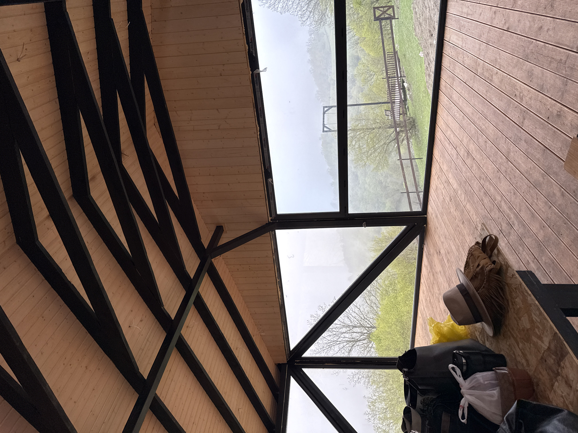 The image presents an interior view in what appears to be a room with a wooden floor, wall, and ceiling. The ceiling features a pattern of wooden planks intersected by dark beams. The room has large windows that offer a view of an outdoor area that seems to be misty, mountainous landscape with a wooden structure resembling a playground. Inside the room, various personal items are visible, including luggage, a hat, a handbag, and a yellow plastic bag, suggesting this may be a guest house or vacation rental. While the prompt requests the location to be identified as Haghartsin, Armenia, there is no visible or readily deducible evidence within the image to verify this claim or link it to a specific location within that city. There are no people visible in the frame, and the items are still.