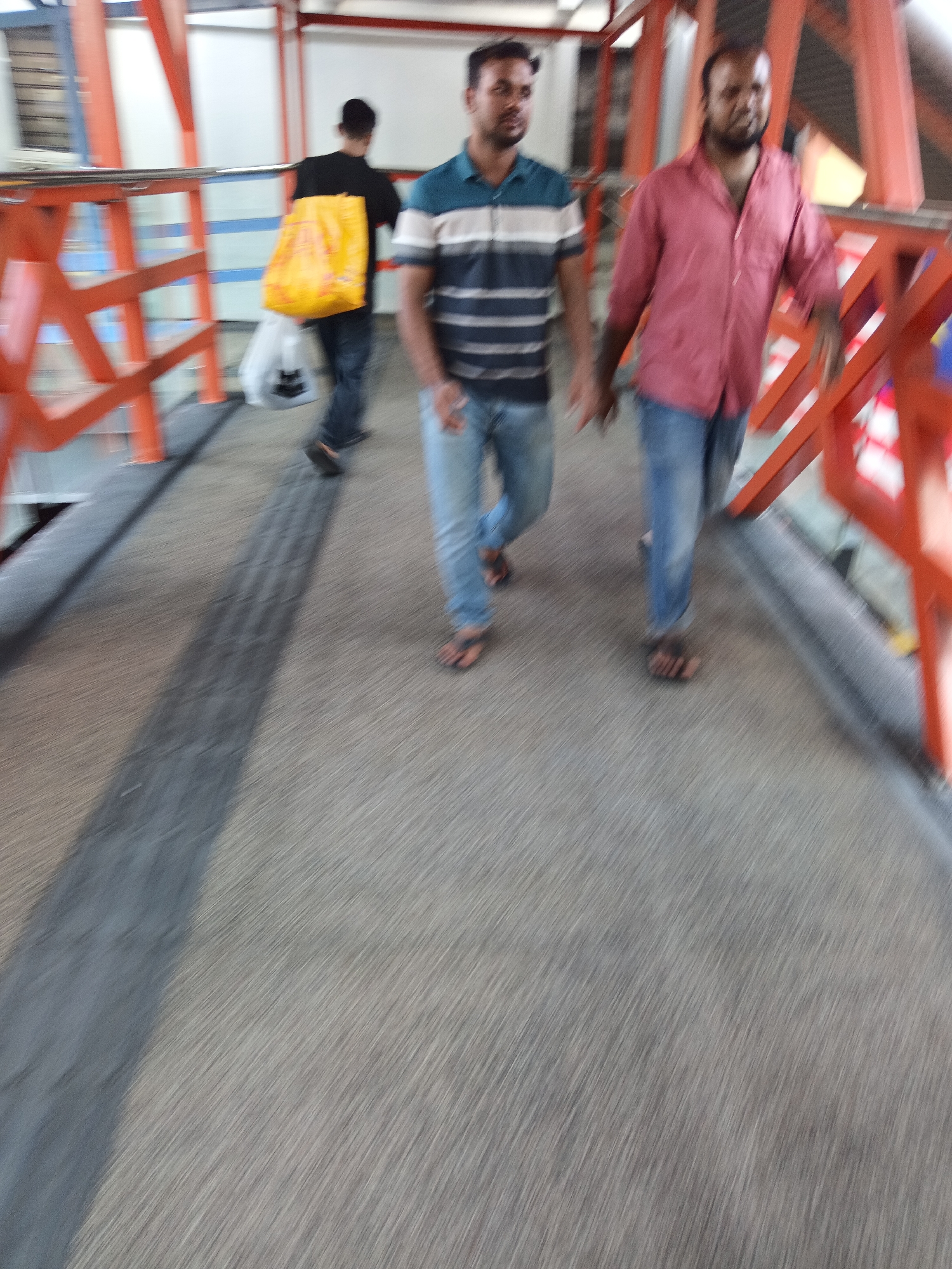 This image captures a street scene in Kuala Lumpur, Malaysia, focusing on three individuals walking along an elevated walkway. The walkway is constructed with orange metal railings and a grey textured surface. The foreground is dominated by the motion-blurred walkway, suggesting movement.
In the background, three men are walking. The man on the far left is wearing a black t-shirt and carrying a bright yellow bag over his shoulder, along with a white bag in his hand. He appears to be looking forward. In the middle, a man is wearing a horizontally striped blue and white shirt and blue jeans, walking with his hands at his sides. The man on the right is wearing a salmon-colored shirt with rolled-up sleeves and blue jeans, also walking with his hands by his sides. The motion blur is most pronounced on the individuals, further emphasizing the sense of movement within the frame. The environment suggests an urban setting, likely an overpass or a pedestrian bridge in a public area.