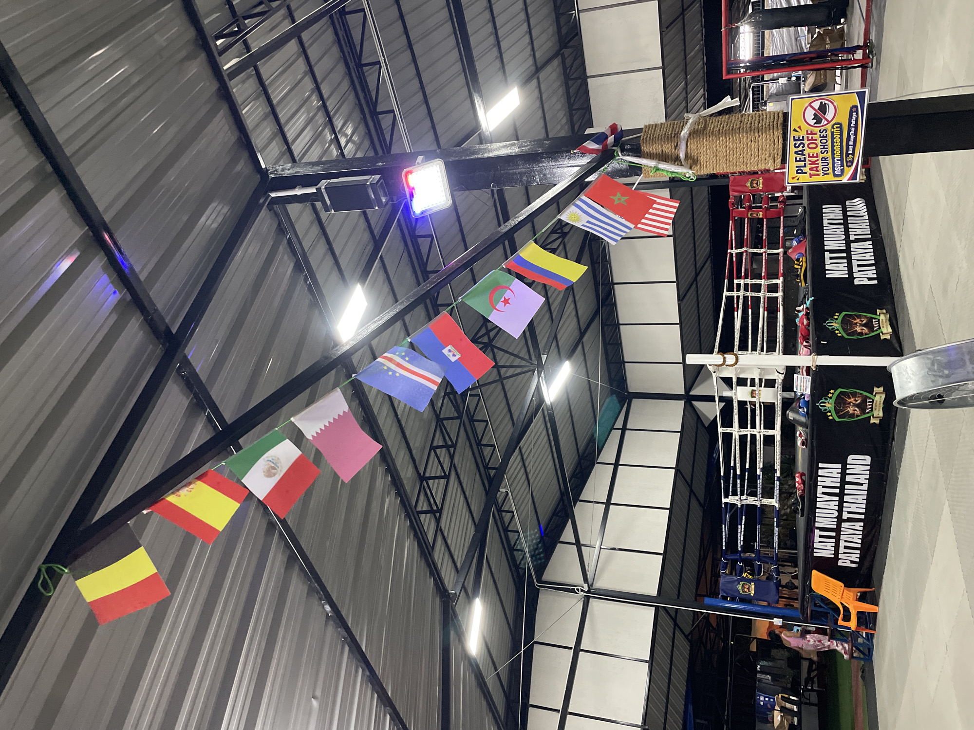 The image shows the interior of a Muay Thai gym in Pattaya City, Thailand, specifically at Natt Muay Thai Pattaya Thailand. The gym features a boxing ring with white ropes and a black skirt displaying the gym's name and logo. Several flags of various countries are strung across the ceiling, suggesting an international clientele or a celebratory atmosphere. A sign near a pole reads "PLEASE TAKE OFF YOUR SHOES" in English and Thai. In the background, there are training equipment like punching bags and mats, as well as a person in pink attire standing near the ring. The overall environment is utilitarian, designed for combat sports training.