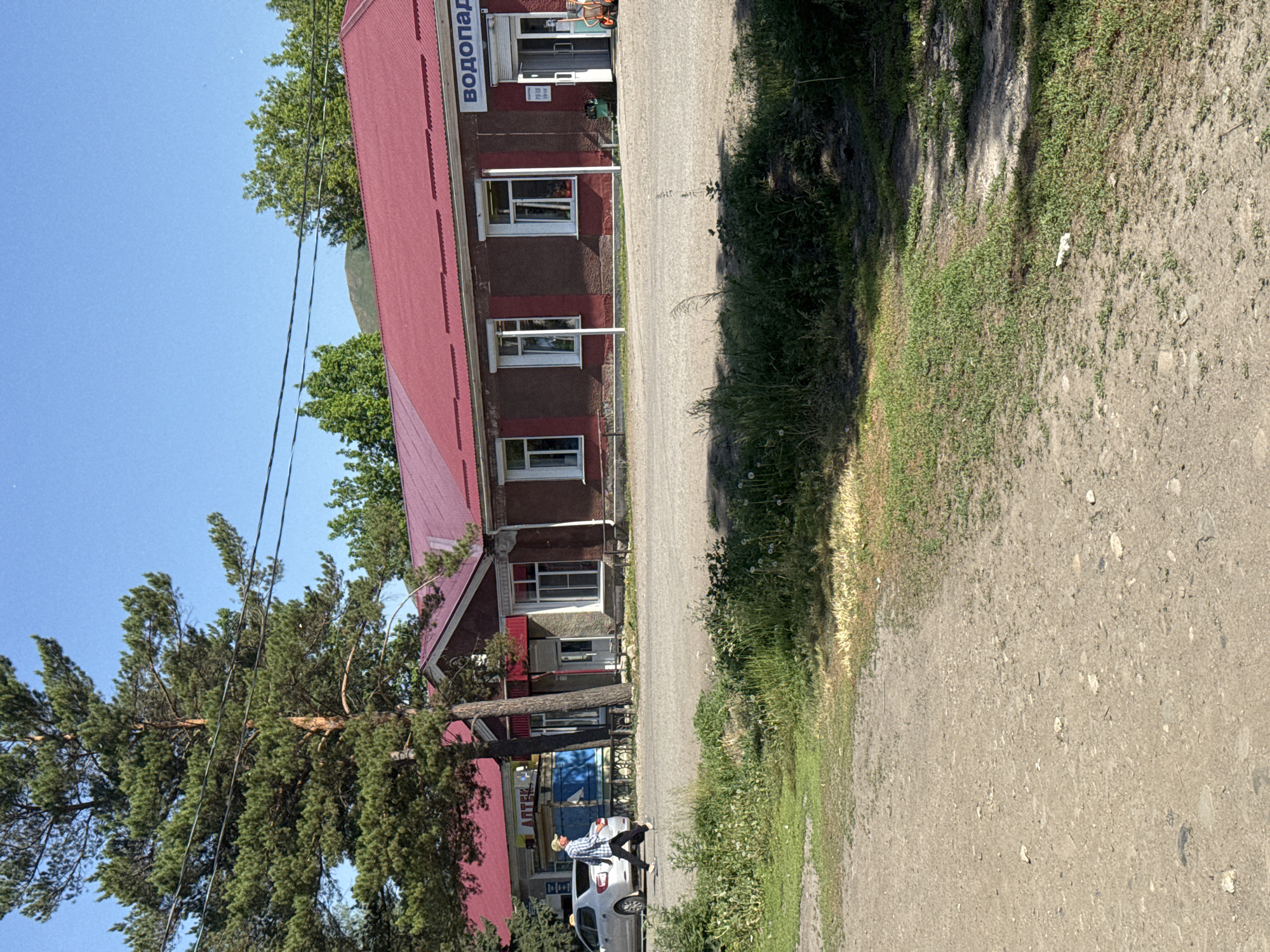 A man in a light-colored cap and plaid shirt walks across a gravel lot in front of a one-story building with a red roof. To the left, a silver sedan is parked. The building has several windows and a sign above the entrance that reads "ВОДОПАД" (Waterfall). To the left of this sign, another sign reads "АПТЕК" (Pharmacy). The scene appears to be in a rural or small town setting, possibly on a sunny day.