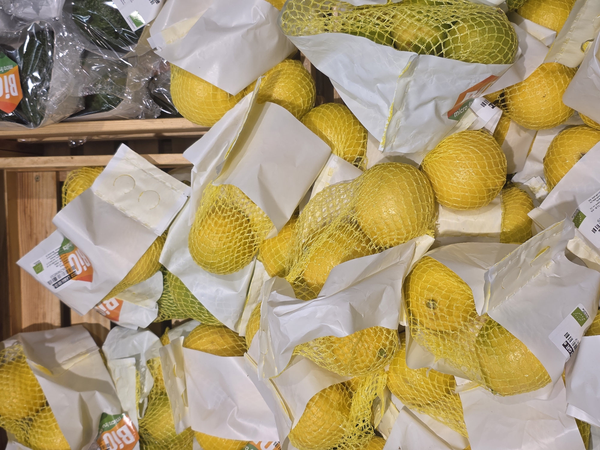 The image shows a close-up, top-down view of a pile of ripe lemons, each encased in a yellow mesh netting and white paper wrapping. The lemons are vibrant yellow, with some showing hints of green. They are loosely piled in what appears to be a display, possibly in a market or grocery store. The background consists of wooden crates and some plastic-wrapped produce, including what looks like avocados. Labels visible on the packaging indicate "Bio" (organic) and some text in a foreign language, possibly Romanian, given the mention of Bucharest. There are no people or animals in the image. The lighting is bright, suggesting an indoor setting.