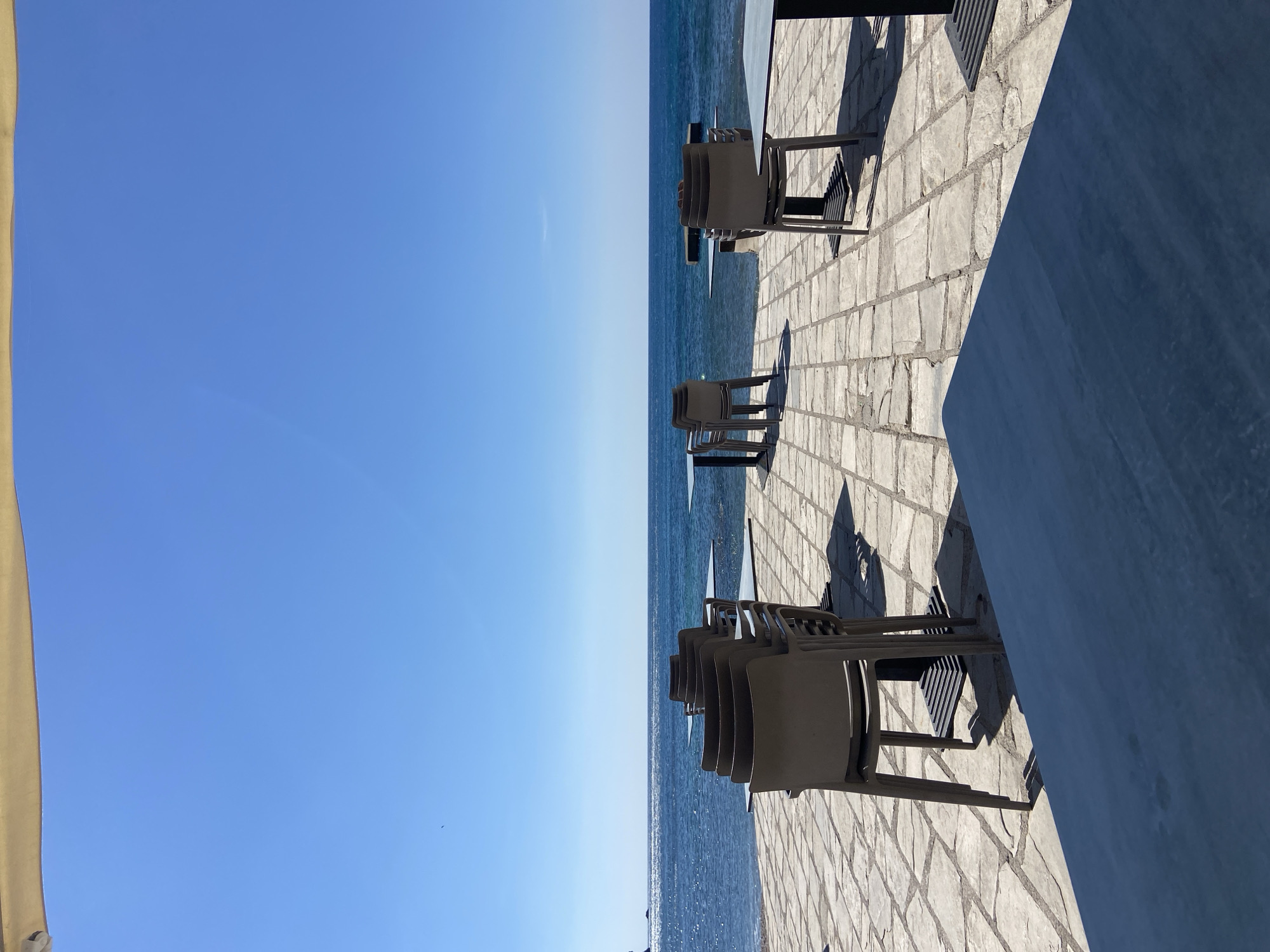 The image shows a view of the Adriatic Sea from an outdoor dining area in Budva, Montenegro. The sky is clear and blue. The sea is a slightly darker shade of blue, with some visible wave patterns. Along the edge of the dining area, several stacks of plastic chairs are placed on a stone-tiled terrace. A table is partially visible in the right foreground. The scene is bathed in sunlight, casting shadows from the furniture.