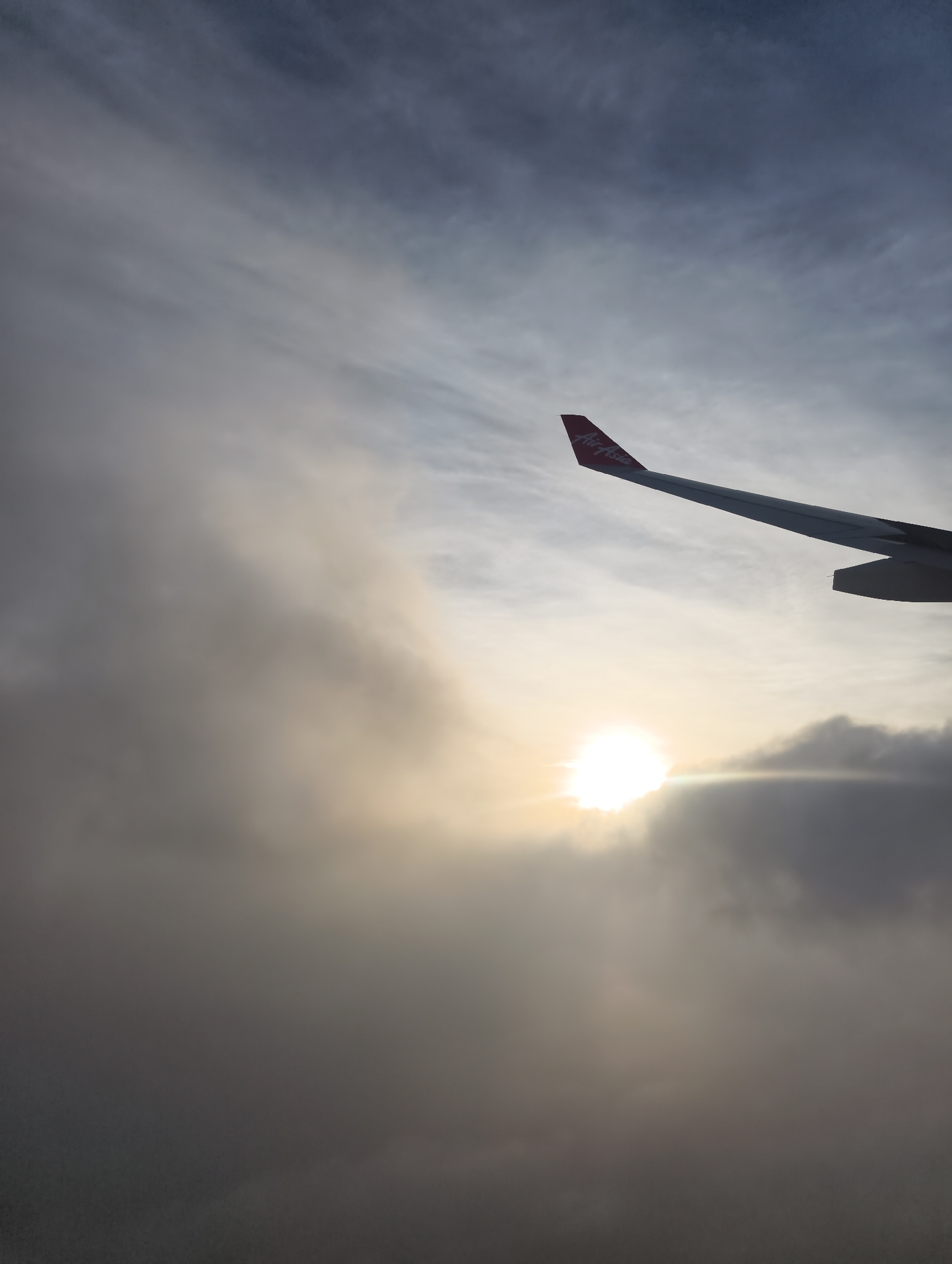 The image shows an airplane wing, with the "Air Asia" logo visible on the winglet. The background consists of cloudy sky with the sun shining through the clouds, creating a bright glare. The location is airborne, above the clouds. The specific city and location are unknown.
