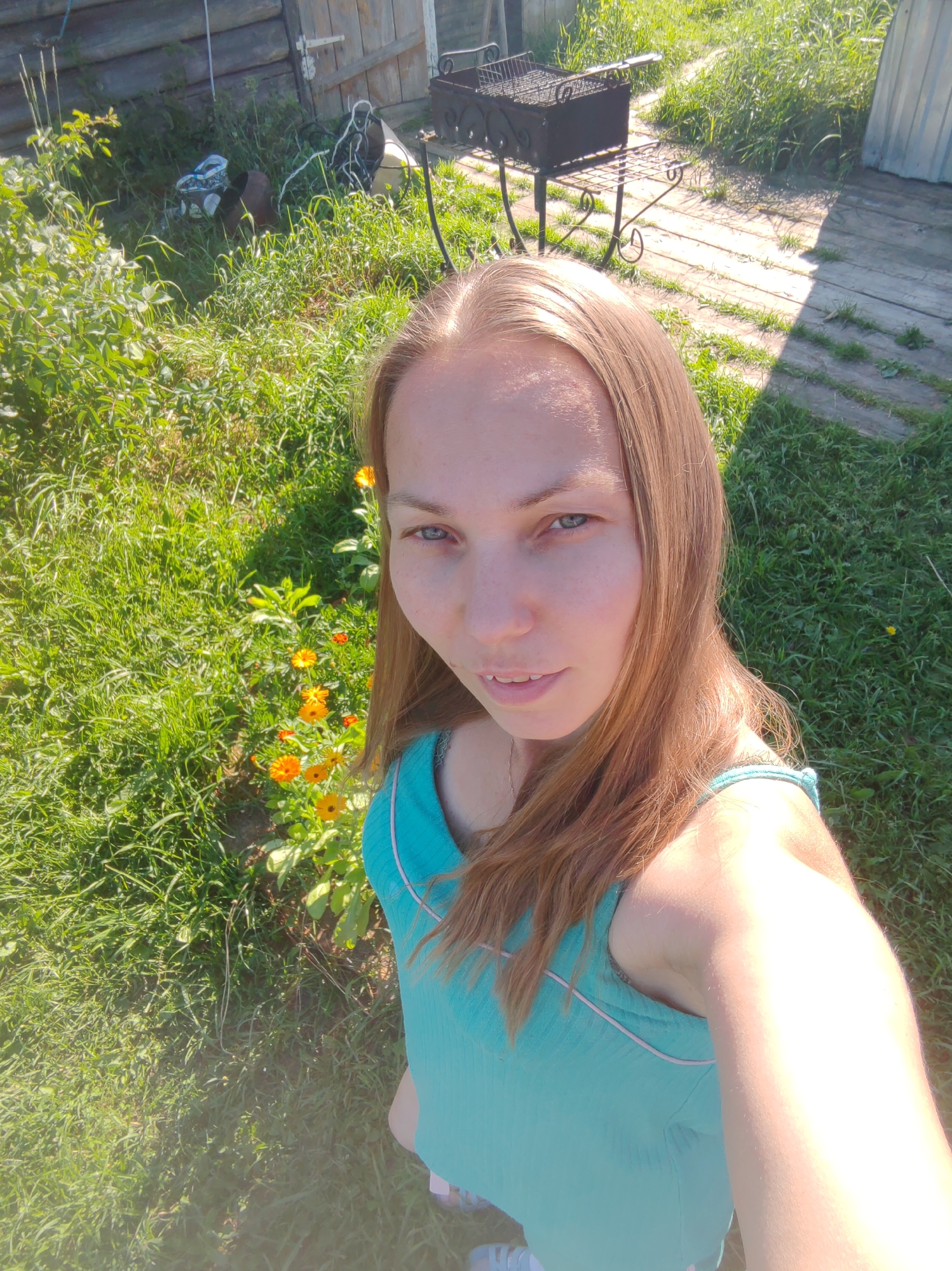 In Чухлэм, Russia, a young woman with long, light brown hair takes a selfie outdoors. She is wearing a teal tank top and is standing in front of a wooden structure, possibly a fence or wall, made of logs. To her right, there is a metal barbecue grill on a stand, and beyond that, a wooden plank walkway. The ground is covered with lush green grass, and in the foreground, a small cluster of orange flowers is visible. The scene appears to be a sunny day in a rural or backyard setting.