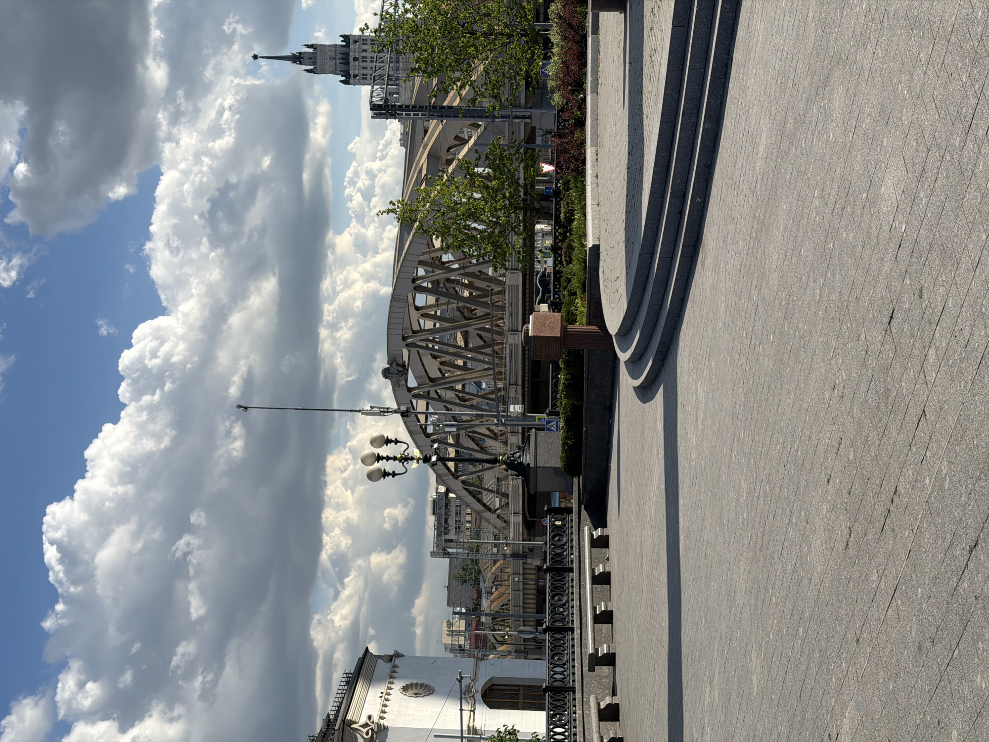 This image captures a vibrant, sunny cityscape along what appears to be a river embankment in Moscow. The perspective is from a paved walkway, looking towards a prominent bridge and city skyline.
In the foreground, a wide, light-grey paved area, possibly granite, with a subtle diamond pattern, occupies the right side of the frame. To its left, a series of curved stone steps ascend, casting long, distinct shadows across the pavement. A small, dark red or brown pedestal or marker is visible near the base of the steps.
The midground is dominated by a large, ornate metal truss bridge, likely a railway bridge, with visible tracks and overhead power lines. Green trees and dense bushes are scattered along the embankment and beneath the bridge structure. A classic black streetlamp with multiple white spherical globes stands to the left of the paved area, near the bridge. A small, triangular road sign with a red border and a black pedestrian crossing symbol is visible among the greenery.
In the background, beyond the bridge, a distinctive tall building with a pointed spire, characteristic of Moscow's Stalinist-era architecture (one of the "Seven Sisters"), stands prominently against the sky. Other city buildings are discernible in the distance.
The sky is a bright blue, filled with large, fluffy white cumulus clouds, indicating clear weather. The strong shadows cast by the steps and other structures suggest bright sunshine, likely during the mid-morning or afternoon. No people are visible, leaving the scene serene and architectural.