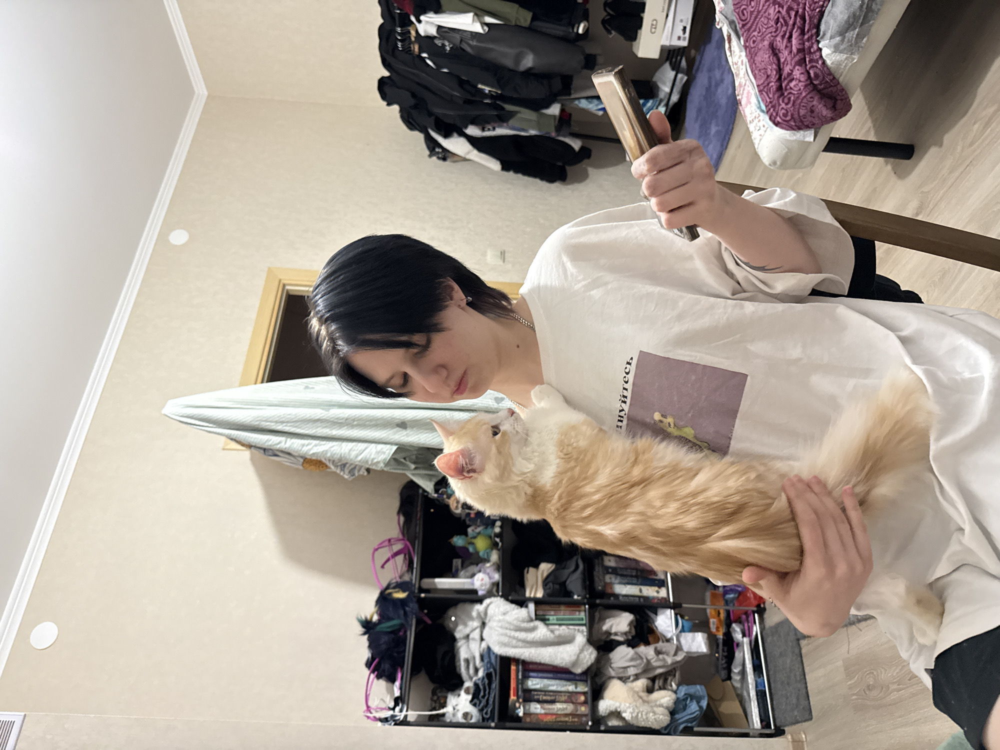 This image captures an indoor residential scene within the city of Saint Petersburg, Russia. A young individual with short, dark hair, wearing a white oversized t-shirt with a graphic print, is seated while holding a long-haired, light orange and cream-colored cat. The cat is positioned on the individual's lap and chest, with its left front paw resting on the individual's left shoulder, and its head turned towards the left. The individual's left arm supports the cat, while their right hand holds a rectangular, dark object, resembling a mobile phone. The individual has small silver hoop earrings, a silver chain necklace, and a dark tattoo on their inner right forearm.
The room features light beige textured wallpaper, white crown molding, and a light wood-patterned floor. In the left background, a black multi-shelf unit is densely filled with books, folded clothing, and miscellaneous items including small plush toys. In the right background, a black garment rack on wheels is heavily laden with dark clothing items on hangers. Below the rack, shoeboxes and a pair of dark shoes are visible. A section of a white bed frame or bench with dark legs is in the lower right foreground, covered by a folded purple patterned blanket. A light blue/white striped fabric hangs over a wooden door frame in the mid-background. A rectangular ventilation vent is visible on the upper left wall, and a white circular fixture is on the ceiling.