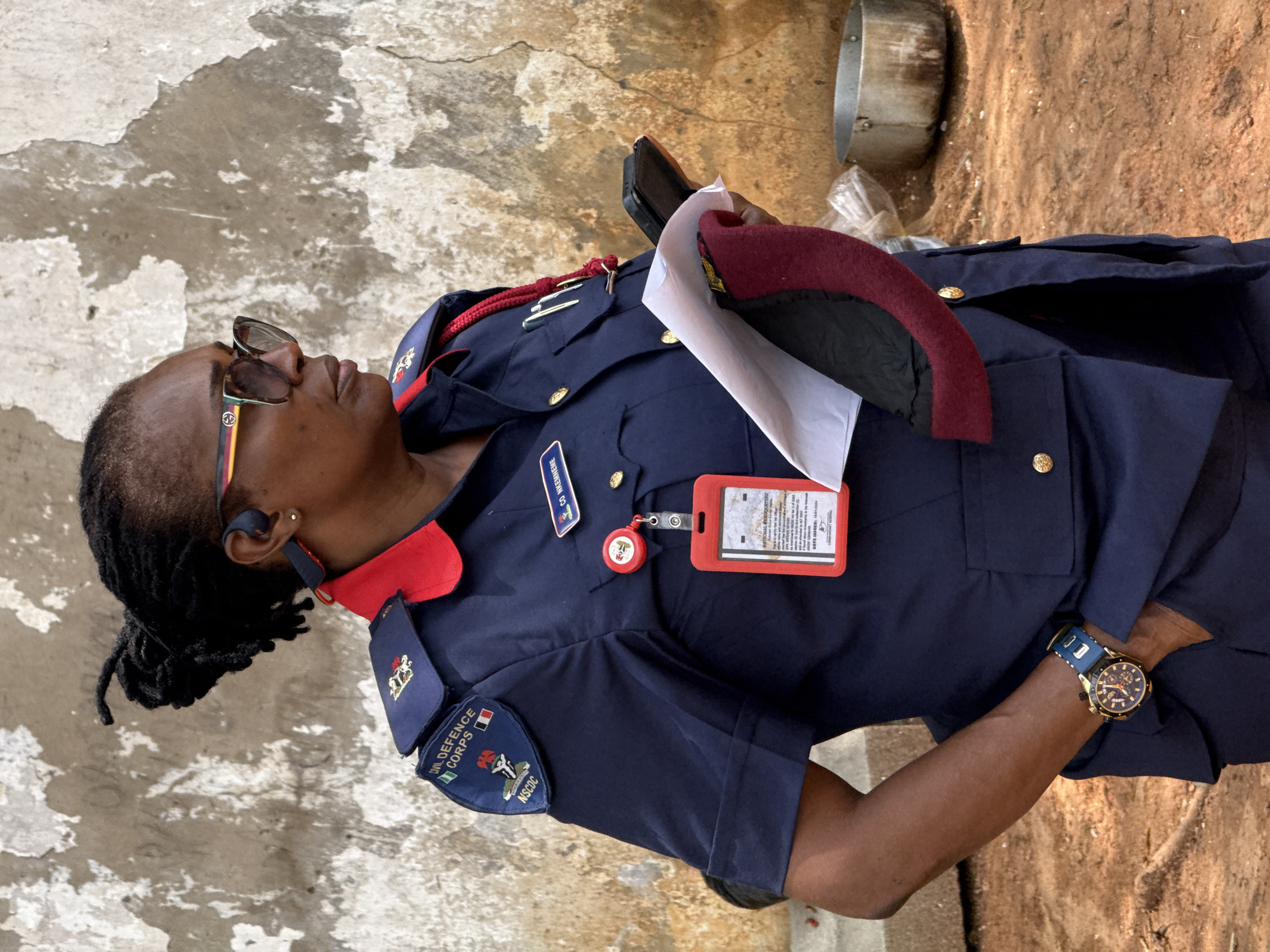 A medium shot shows a woman wearing a blue uniform with red trim, glasses, and a blue watch. The uniform has a patch on the left sleeve that reads "Civil Defence Corps" and shows the Nigerian flag, and also has a name tag visible on the right side of her chest. She is also wearing earbuds, has her hair up in dreadlocks, and is holding a maroon beret with a white piece of paper underneath. The location is likely the city of Abuja, Nigeria, given the patch on the uniform and the name tag. The background shows a textured wall and ground with some scattered debris.