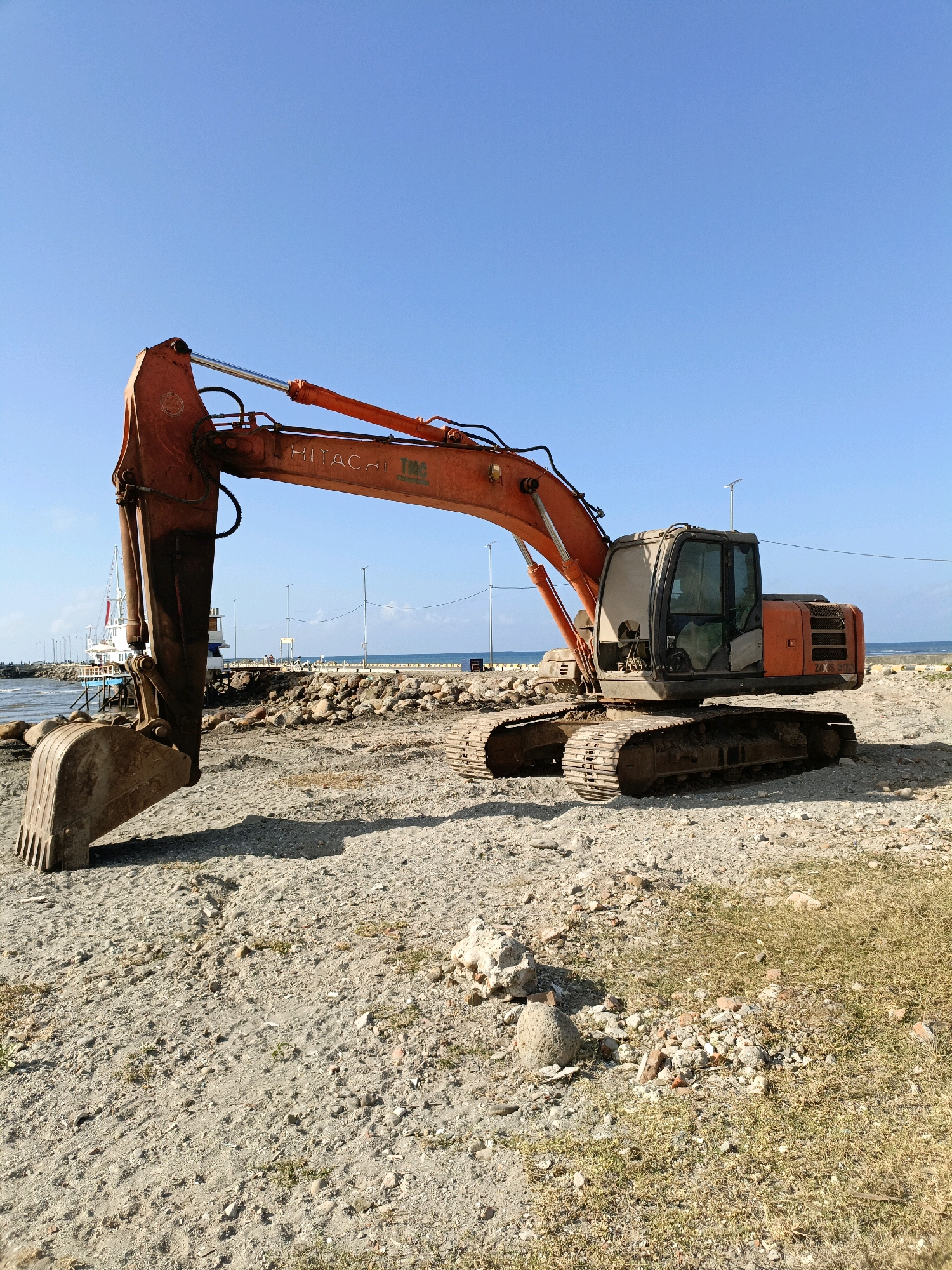 An orange Hitachi excavator is parked on a sandy beach next to a body of water, likely the sea, under a clear blue sky. The excavator is in the foreground, taking up the majority of the image. It is a large piece of construction equipment with tracks, a boom, and a bucket. The excavator's cab is visible, and the brand name "HITACHI" is clearly displayed on its boom.
In the background, there is a pier or jetty extending into the water. On the pier, there are some buildings and structures, including what appears to be a white boat or ship. Several lampposts are also visible along the pier. The water is calm, and there are some rocks and debris scattered on the beach in front of the excavator. The overall impression is of a construction site near the coast.
Given the presence of a Hitachi excavator and its likely use in construction or infrastructure projects, and considering the port-like structures in the background, this scene could plausibly be located in the vicinity of the port of Bulukumba, Indonesia, which is known for its shipbuilding and maritime activities.