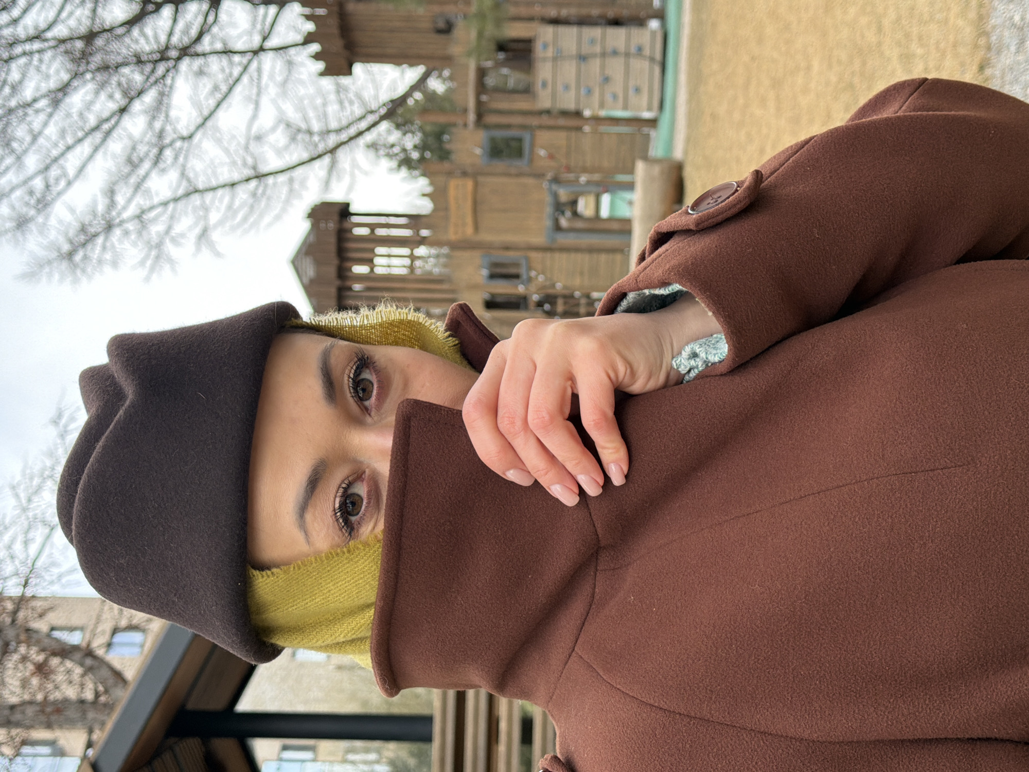 A woman stands outdoors, partially concealed by a brown coat pulled up over her mouth and nose. She wears a dark brown hat and a yellow under-layer around her face. Her eyes are visible, and one hand is positioned near her face, holding the coat. In the background, slightly blurred, is a wooden playground structure, situated in what appears to be a park or public area. The setting seems to be urban. Given the request for location details, the scene appears to be a park location of "Gorky Central Park" in Krasnoyarsk, Russia.