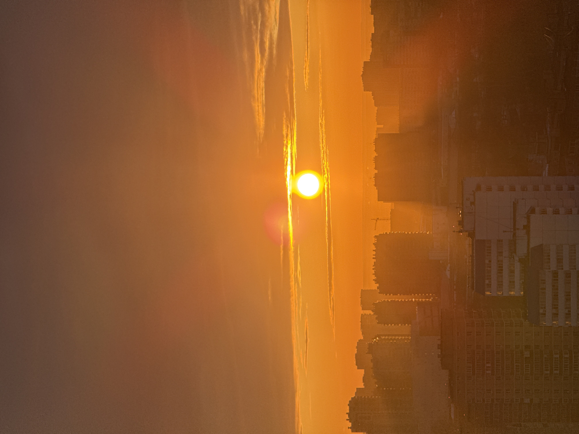 An urban panoramic view of Perm, Russia, is captured during either sunrise or sunset, characterized by an intensely warm color palette dominated by orange and yellow hues. The upper portion of the frame displays a gradient sky, transitioning from a reddish-brown at the zenith to a vibrant orange and yellow near the horizon. A bright, circular, white-yellow sun, exhibiting a distinct lens flare, is centrally positioned horizontally and approximately one-third down from the top edge. Sparse, elongated cloud formations are visible, particularly around the sun.
The lower two-thirds of the image present a dense cityscape, largely silhouetted against the brilliant sky. In the immediate foreground, partially visible multi-story buildings include a lighter-colored structure on the left with numerous visible windows, and a darker, rectangular building to its right. Behind these, a prominent and dense line of uniformly dark, multi-story urban buildings forms a continuous silhouette across the midground. Further towards the horizon, additional layers of more indistinct building silhouettes extend, appearing hazy. A solitary construction crane is faintly discernible on the far right horizon, silhouetted against the sky. No individuals or specific actions are visible. The environment is a densely developed urban setting, viewed from an elevated vantage point.