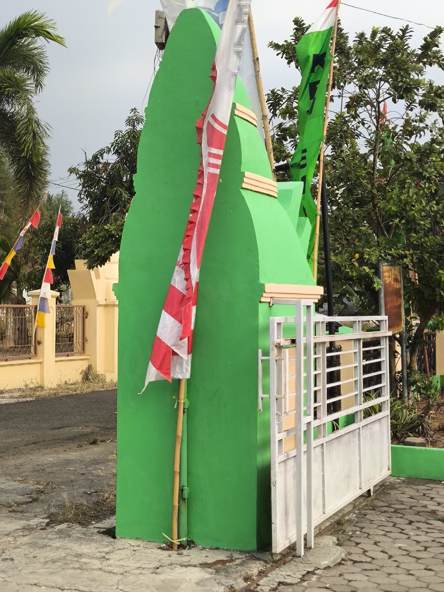 In the city of Cilegon, Indonesia, a bright green arched structure stands prominently near a road. The structure is adorned with several flags, including a red and white Indonesian flag on a bamboo pole, and other flags with green and black designs. To the right of the green structure, a white metal gate is visible, leading to an area with trees and other vegetation. In the background, a light yellow building with a fence can be seen, and the sky is overcast. The overall scene suggests a cultural or religious site decorated for an event or celebration.