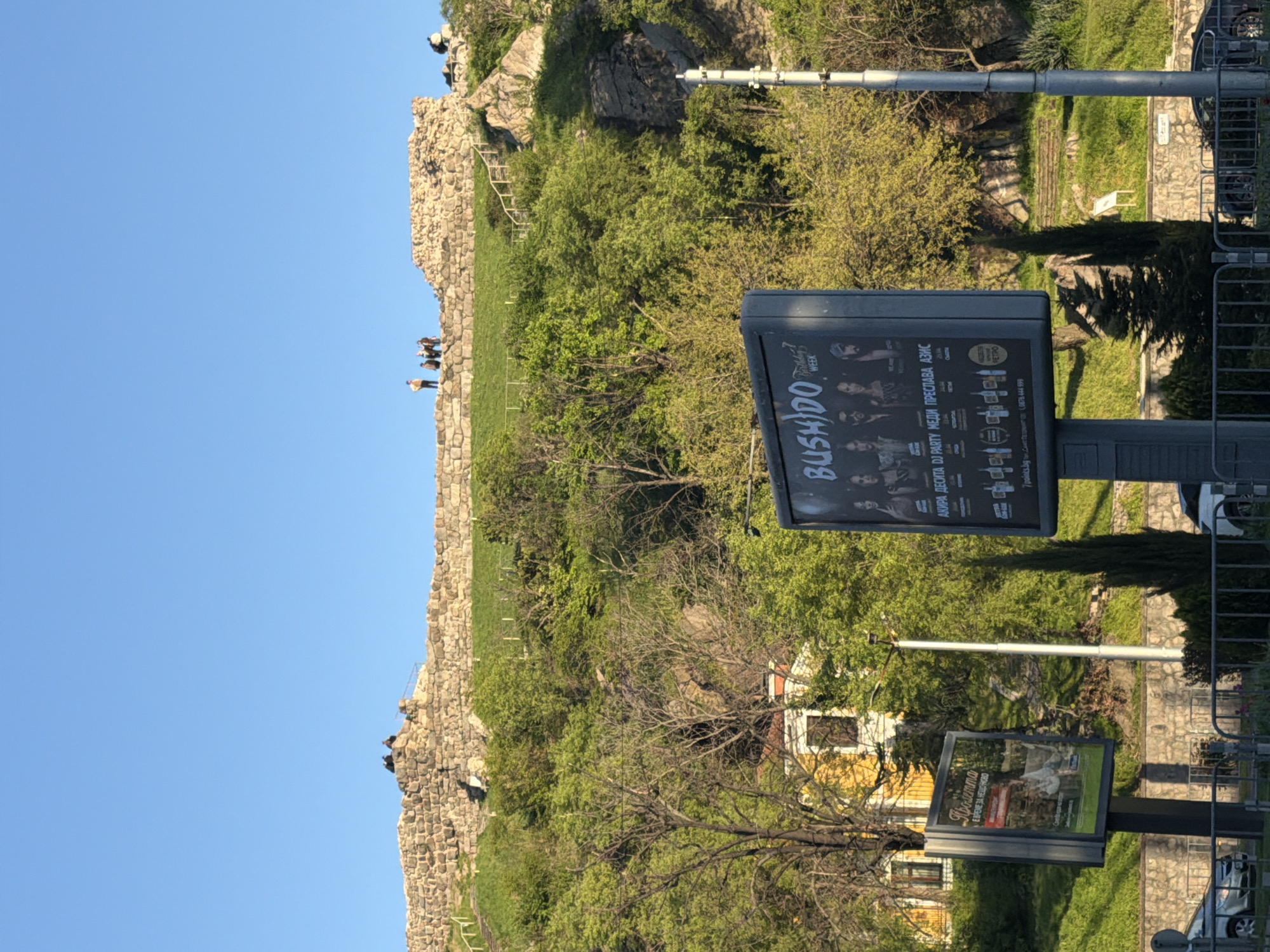 The image depicts an outdoor scene in Plovdiv, Bulgaria, on a clear, sunny day, likely in spring or early summer given the vibrant green foliage. The prominent feature on the left is a substantial ancient stone wall, characteristic of historical fortifications found on Plovdiv's hills. Several people are visible atop this wall, seemingly enjoying the view from the elevated position, with some individuals also further up on the rocky, tree-covered hillside behind it.
In the foreground to the right, two large vertical billboards are displayed on poles. The upper, larger billboard advertises "BUSHIDO WEEK" and lists various Bulgarian music artists and event types, including "AKIRA ДЕСИТА DJ PARTY МЕДИ ПРЕСЛАВА АЗИС." It also provides contact details: "7 points.bg" and a phone number "0676 444 999." The lower billboard features the text "Пролетта" (Spring) and "ВРЕМЕ ЗА НЕЩО НОВО" (Time for Something New), suggesting a seasonal theme.
Behind the billboards, amidst numerous trees and shrubs, part of a yellow building with white window frames is visible. Metal railings and glimpses of parked cars indicate a public street or pathway in the foreground, blending the ancient historical elements with modern urban infrastructure under a bright blue sky. The overall impression is one of a lively public space where visitors engage with both the historical landscape and contemporary cultural advertisements.