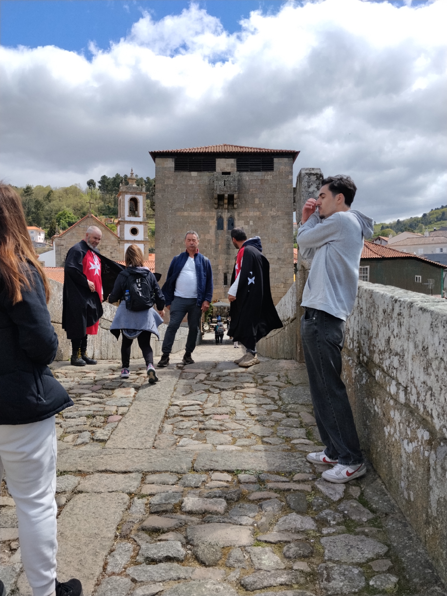 Captured in Tarouca, Portugal, the scene unfolds on a cobblestone bridge under a sky dominated by cumulus clouds. A young man, wearing a grey hoodie and dark jeans with white and red sneakers, leans against the stone bridge wall, his hands near his face. Moving inward from left to right, a woman with long brown hair faces away from the camera wearing white pants and a black jacket. Further in the bridge, another woman with a backpack walks by an older man dressed in a black cape adorned with a white cross; another man in a blue jacket stands nearby. The background features a stone tower with small openings and a church steeple beyond. In the distance, rooftops and hillside foliage are visible.