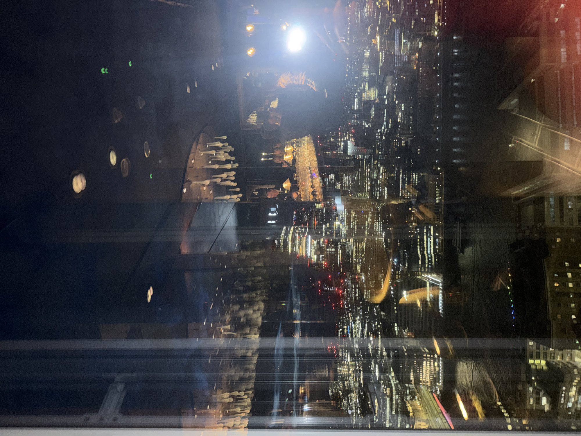 This is a photograph taken from a high vantage point, likely a restaurant or bar with large windows, overlooking a cityscape at night. The scene is a reflection on glass, showing both the interior of the establishment and the exterior view.
In the interior, a bar area is visible with a bartender and patrons. There are rows of glassware on the bar, illuminated by overhead lights and possibly spotlights. A decorative chandelier with multiple teardrop-shaped elements hangs from the ceiling. Some ceiling lights are visible, appearing as blurred circles. There are also some green lights in the upper right portion of the interior reflection, possibly emergency exit signs or decorative elements.
The exterior view is a sprawling urban landscape of a major city at night, consistent with London, United Kingdom. Numerous buildings with illuminated windows create a dense network of lights. A river, likely the River Thames, is visible in the foreground, with lights reflecting on its surface. Boats are present on the river. Bridges and roadways are also discernible within the city. The overall impression is one of a vibrant, bustling metropolis seen from above. The time of day is clearly night, with the darkness accentuating the artificial lights of the city and the interior. The photograph is characterized by reflections and the interplay between the indoor and outdoor environments.