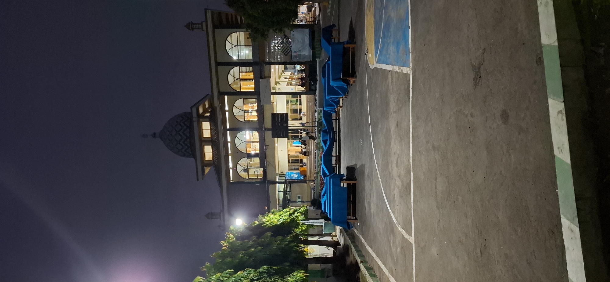 An outdoor, low-angle, nighttime photograph captures a mosque and surrounding courtyard in the city of Wonorejo, Indonesia. The mosque, with its prominent dome, is illuminated from within, casting warm light through arched windows. In the foreground, a paved courtyard, marked with white lines suggesting a sports court, is visible. Several tables draped with blue cloths are arranged across the courtyard. The environment appears to be a public gathering space, possibly for an event or function, given the covered tables. Trees line the left side of the image, and the sky is dark with some atmospheric haze. A few individuals can be seen in the background, near the entrance of the mosque. The overall scene is quiet and dimly lit, suggesting evening or night hours.
