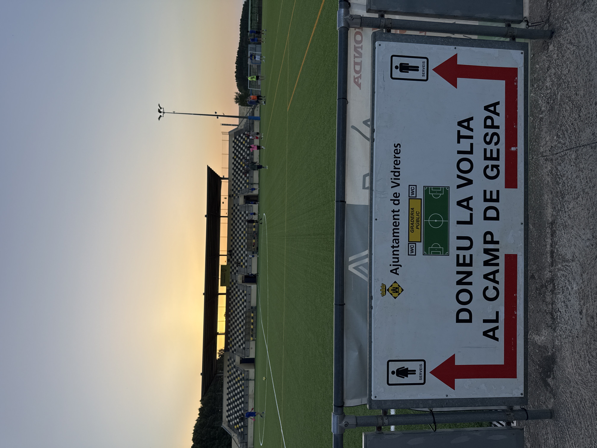 The image depicts a view of a football field at sunset in Vidreres, Spain. In the foreground, a white sign with black text and graphics is prominently displayed. The sign reads "AJUNTAMENT DE VIDRERES" (Town Hall of Vidreres), and below that, "DONEU LA VOLTA AL CAMP DE GESPA" which translates to "Go around the grass field." The sign also indicates "SERVEIS" (Services) with icons for a women's restroom on the left and a men's restroom on the right, each accompanied by a red arrow pointing left and right respectively. The football field itself is green artificial turf, marked with white lines. In the background, a stadium with tiered seating is visible, with the setting sun casting a warm glow across the sky and illuminating the stadium structure. Several individuals are scattered across the field, some appearing to be players, while others are spectating or observing. The overall scene suggests a public sporting facility in use during the evening hours.
