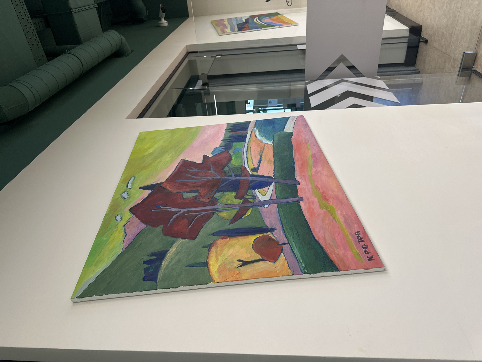 A close-up shot shows a painting displayed in what appears to be a modern art museum in Budva, Montenegro. The painting is an abstract landscape rendered in vibrant colors like green, red, blue, and pink. The artist's signature, "K. POTOJOB," is visible in the lower right corner. In the background is a glass railing, an exhibit with a white chevron and a white object, and an upper floor where another painting is visible. The architectural features suggest an indoor setting with modern design elements. A green industrial pipe is mounted to the ceiling and runs parallel to the painting.