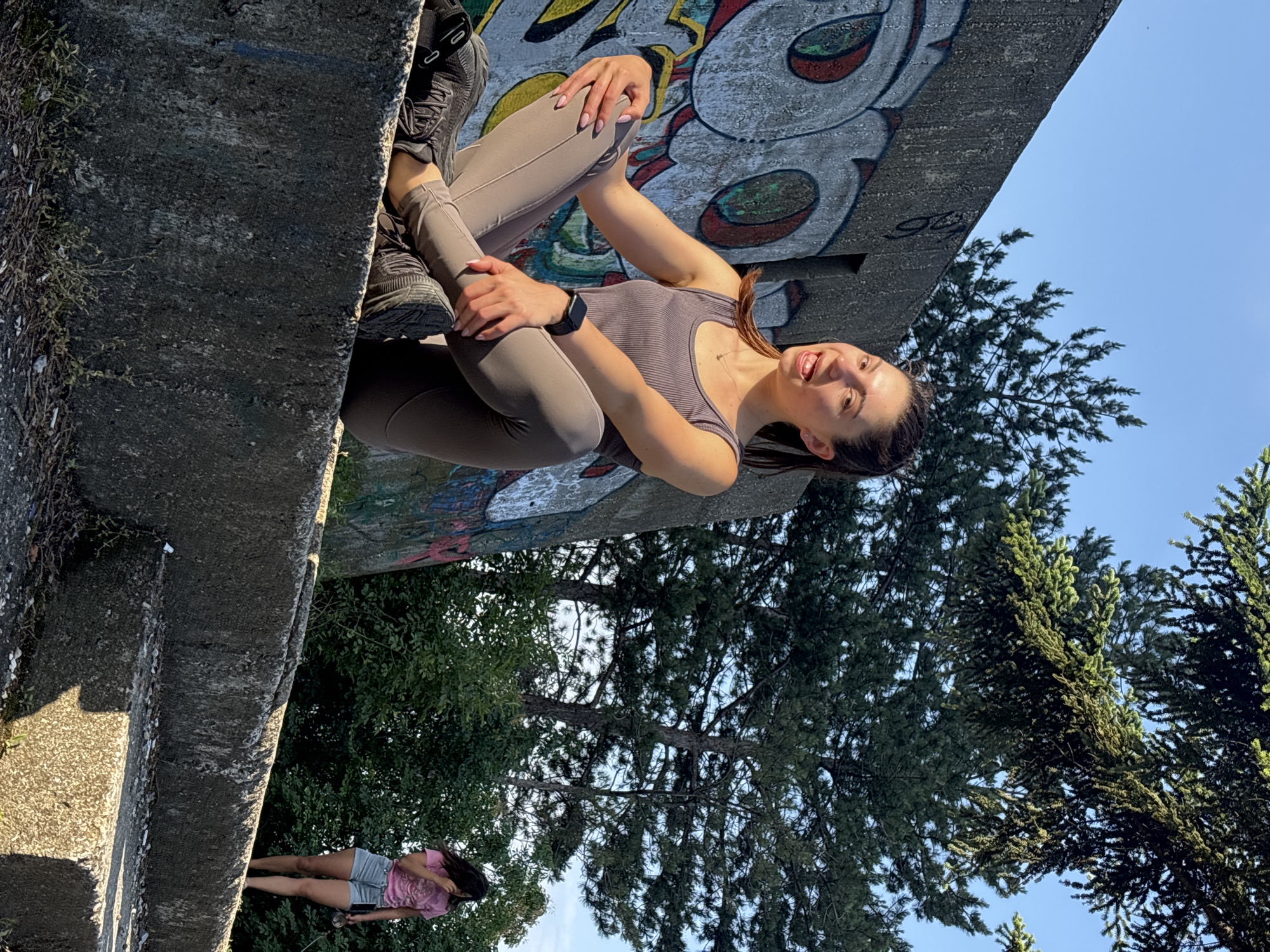 The image, set in Varna, Bulgaria, captures an outdoor scene under a bright, clear blue sky during the daytime. The main subject is a smiling young woman, posing for the camera while seated on a robust concrete ledge. She has dark hair, pulled back, and is dressed in a gray ribbed tank top, matching gray leggings, and dark athletic shoes. A smartwatch is visible on her left wrist, and her fingernails are painted a light shade. She looks up towards the camera with a broad smile, her hands resting on her bent knees.
Behind and above the woman, a concrete wall is decorated with colorful graffiti, showcasing abstract shapes, stylized eye-like elements, and some faint, illegible letter-like forms, including what appears to be "GG." Below the concrete ledge and in the background, a dense growth of green trees, comprising both conifers and deciduous species, suggests a natural or park-like environment.
Further in the background, below the main concrete structure, a second person is partially visible, standing amidst the trees. This individual is wearing a light pink t-shirt and shorts and appears to be holding an object. The overall mood of the image is cheerful and relaxed, bathed in bright sunlight.