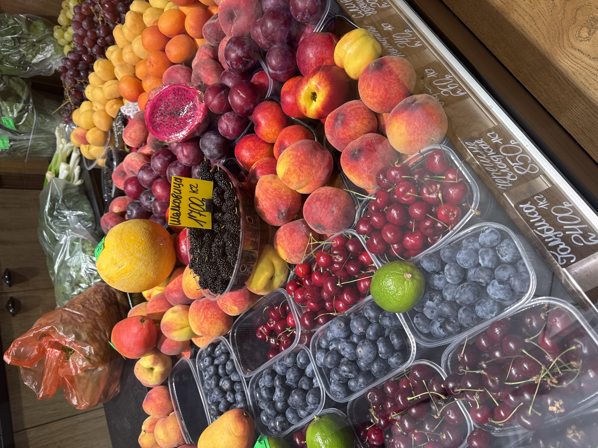 This image captures a vibrant and extensive display of fresh fruits, likely situated in an indoor market or a grocery store in Saint Petersburg, Russia. The scene is filled with an abundant and colorful assortment of produce, neatly arranged in rows and clear plastic containers, some protected by a glass display case.
The subjects include a wide variety of fruits: green and purple grapes, golden apricots, yellow and red peaches, nectarines, and dark plums. An exotic pink dragon fruit, a bright yellow cantaloupe melon, and a partially visible mango add to the diversity. Various berries are prominent, with black mulberries, bright red cherries, and dark blue blueberries meticulously arranged in transparent plastic tubs. Limes are also interspersed among the berries. Green leafy vegetables in plastic bags are visible on the left side of the frame, indicating a broader produce section. The display case features a wooden frame along its top edge.
The activity taking place is the retail sale of fresh fruits. The display is well-lit, highlighting the natural colors and freshness of the produce, suggesting a daytime setting or a brightly lit interior.
Notable details include handwritten price tags, predominantly in Russian, indicating the type of fruit and its price per kilogram in rubles. Visible text on the tags includes:
- A yellow tag for "Шовковиця" (Mulberry) with a price of "170.0 кг" (170.0 Rubles per kilogram).
- White tags under the glass displaying:
- "Нектарин" (Nectarine) at "240 ₽/кг".
- "Персик" (Peach) at "250 ₽/кг".
- "Черешня" (Cherry) at "850 ₽/кг".
- "Алича" (Cherry Plum) at "100 ₽/кг".
- Partially visible tags for "Слива" (Plum) at "350 ₽/кг" and "Виноград" (Grape) at "380 ₽/кг".
No people are visible in the image, allowing full focus on the colorful and organized fruit display.