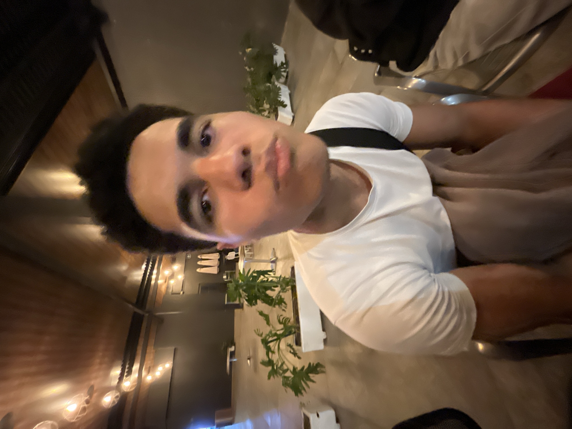 A young man is taking a selfie indoors. He has curly dark hair, dark eyes, and is wearing a white t-shirt with a black strap or bag strap across his shoulder. The environment appears to be a restaurant or bar, featuring plants in planters, a brick wall, and string lights. The precise location within San Bernardino Tlaxcalancingo, Mexico, cannot be determined from this single image.