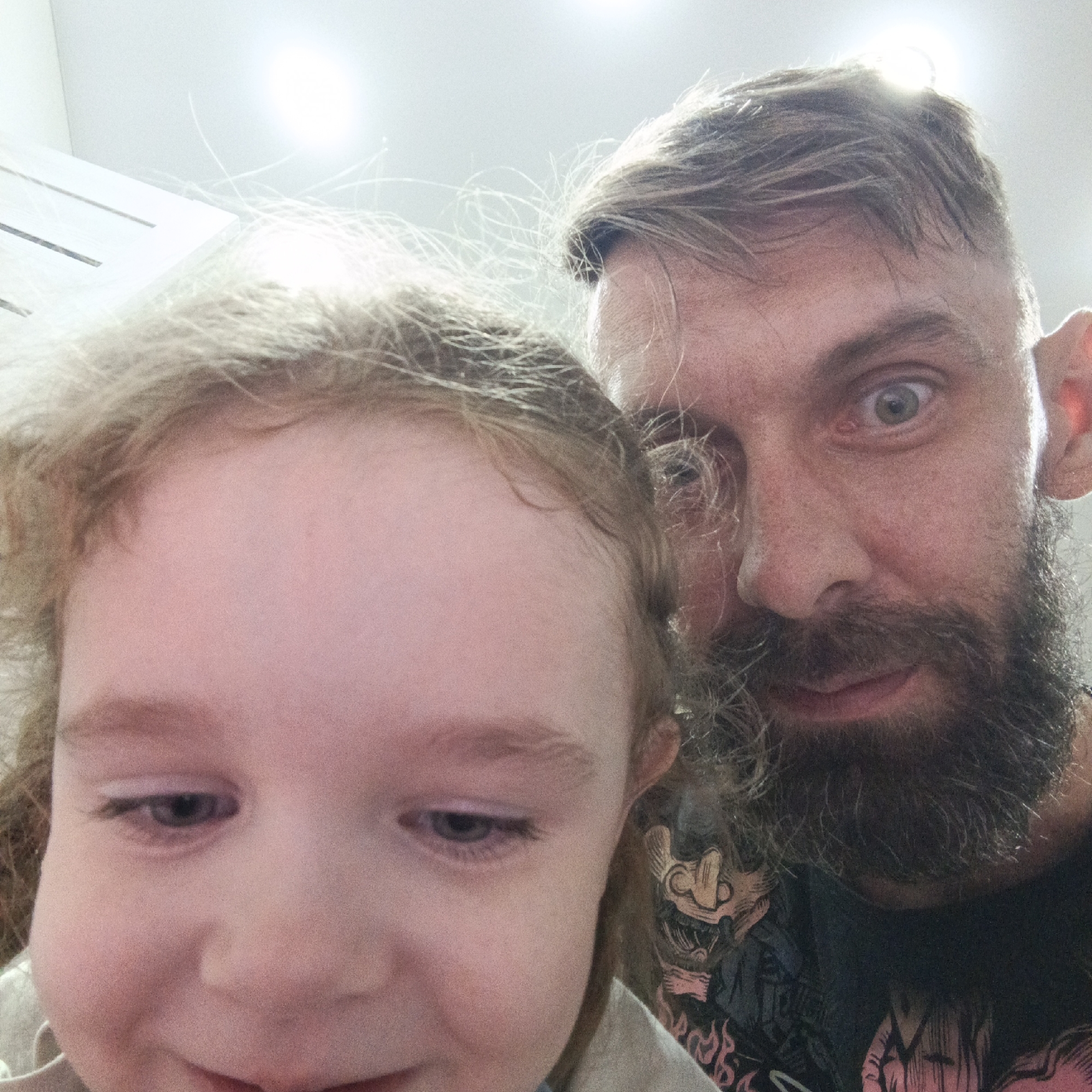 This image shows a selfie taken at a close range, featuring a man and a young child. The man is on the right side of the frame and has a beard, short hair, and wide-eyed expression. A small portion of his t-shirt is visible, which has a graphic design. The child is positioned on the left side of the frame with a downward gaze. The environment is indoors with bright overhead lighting. There is no indication the photo was taken in Марковский, Russia.