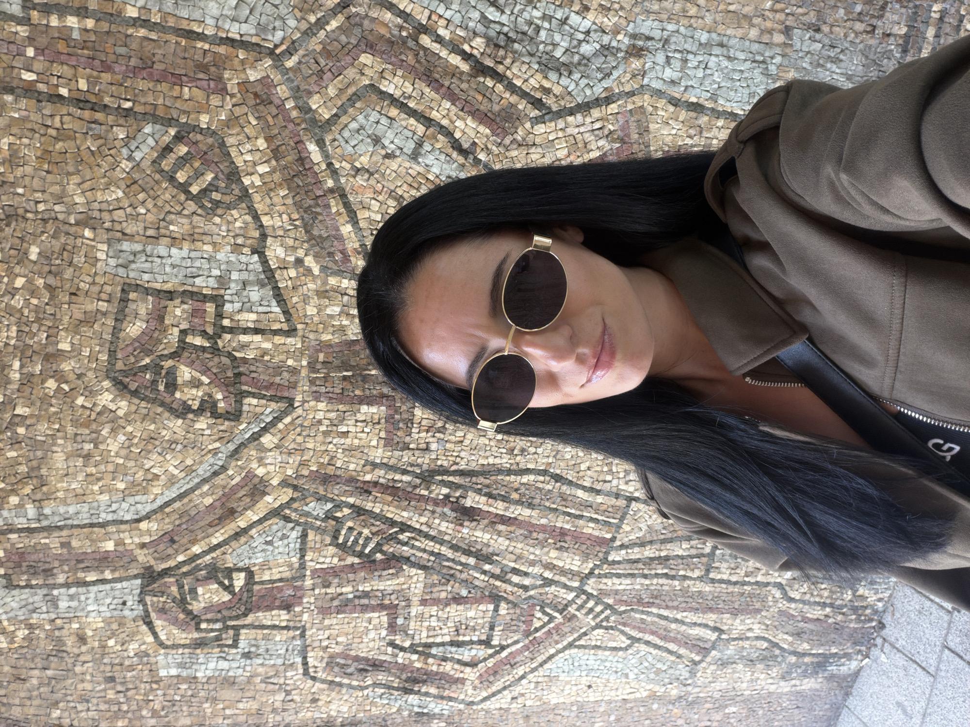 A woman with black hair and sunglasses stands in front of a large mosaic in Pleven, Bulgaria. The mosaic is composed of small, irregularly shaped tiles in earthy tones. The mosaic depicts figures and geometric patterns. The woman is wearing a brown jacket and has a black purse or bag. The woman's features are sharp, and she has a neutral expression. The environment is outdoors.