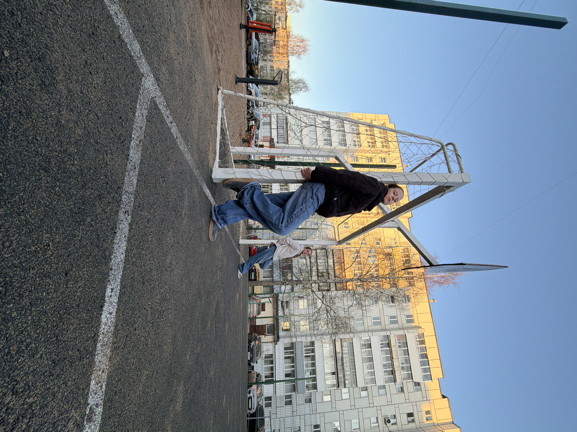 In a public space in Chaikovsky, Russia, two people are interacting with a soccer goal on a paved surface marked with white lines. One person, wearing a black jacket and blue jeans, is hanging from the goal, suspended horizontally between the goalposts. Another person, wearing a light-colored jacket and blue jeans, is standing nearby, partially obscured. In the background, there are apartment buildings, bare trees, and parked cars. The sky is clear and blue.