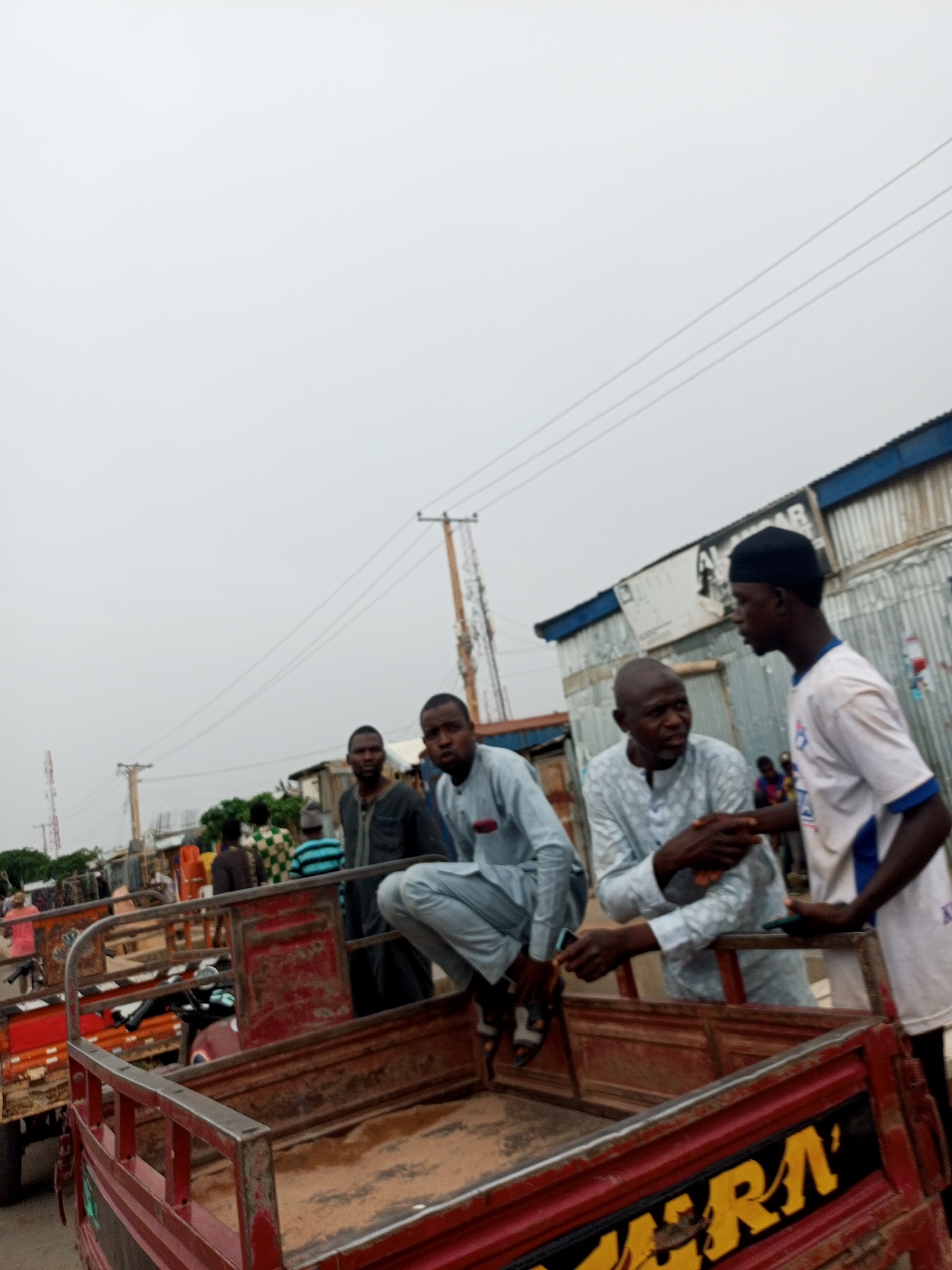 The image shows a group of men in an urban area of Nigeria, possibly Tempure, based on the implied location. Four men are interacting near the back of several flatbed carts. One man is squatting on the cart's edge wearing a light blue suit, another man is standing in a white t-shirt and black cap, and a third man is standing next to him wearing a light blue patterned shirt. The fourth man is further back, also wearing a dark shirt or jacket. The men appear to be in conversation.
The carts are predominantly red with some rust and wear. One cart has lettering visible, reading "ERRA".
The background includes a building made of corrugated metal, power lines, utility poles, and other indistinct figures. The sky is overcast. The scene appears to be a street or market area.
