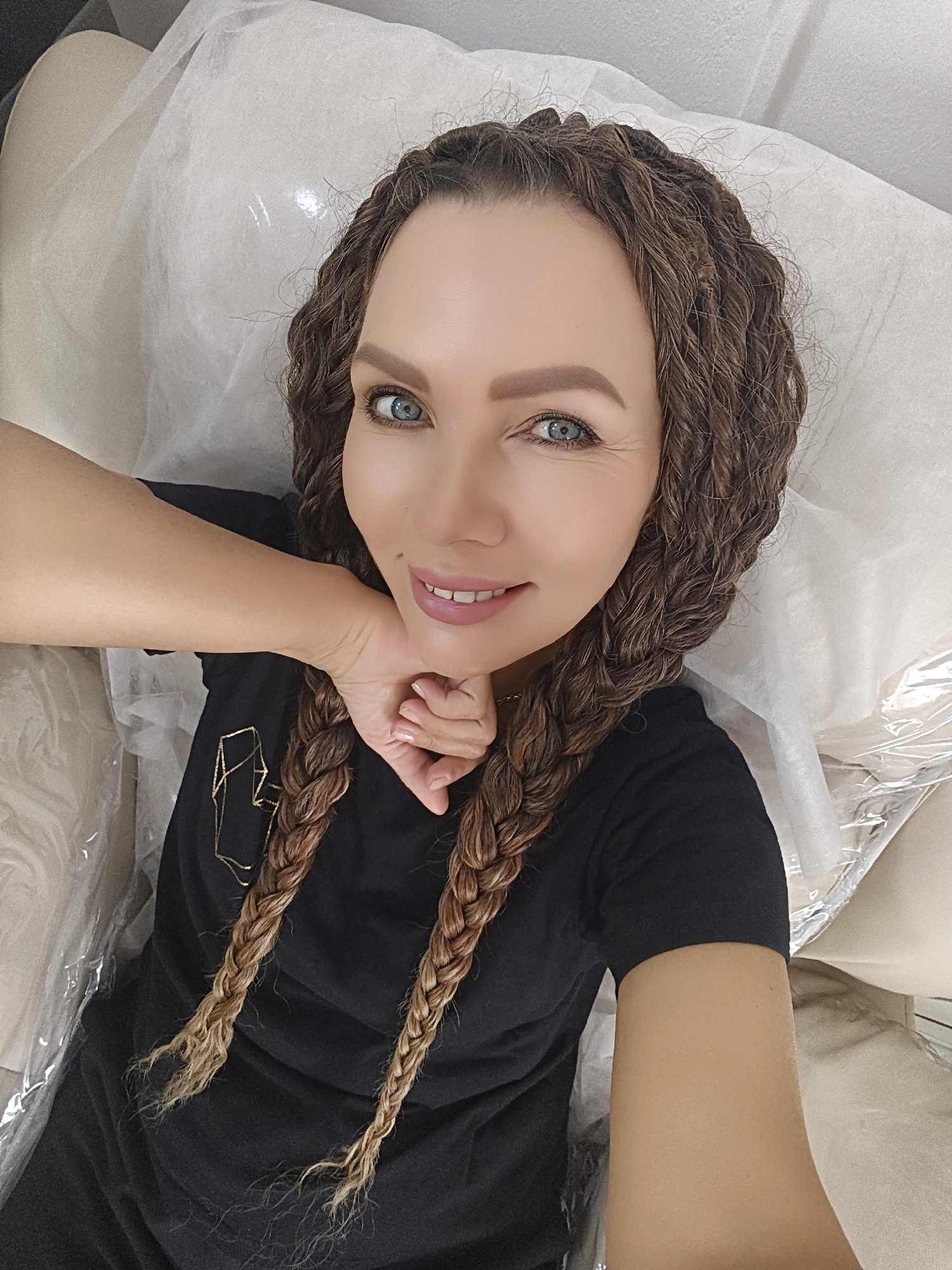 A woman is featured in a close-up, high-angle selfie, smiling gently while reclining on what appears to be a treatment bed or chair. Her head is slightly tilted, and her right hand rests delicately under her chin. She has light skin, striking blue-green eyes, and her light brown hair is styled in numerous small, curly braids, some of which merge into two longer braids that fall over her shoulders with lighter, possibly ombre, ends. Her eyebrows are well-defined, and she wears subtle makeup, including pink lipstick.
She is dressed in a black short-sleeved t-shirt that features a gold, abstract, geometric design on the front. The background consists of a light-colored surface, likely upholstery, covered with a white, disposable sheet or paper and sections of clear plastic sheeting, suggesting a hygienic environment such as a salon, spa, or clinic. The woman appears calm, relaxed, and content, engaging directly with the camera. The lighting is soft and even, indicating an indoor setting. There is no discernible text other than the abstract design on her shirt.