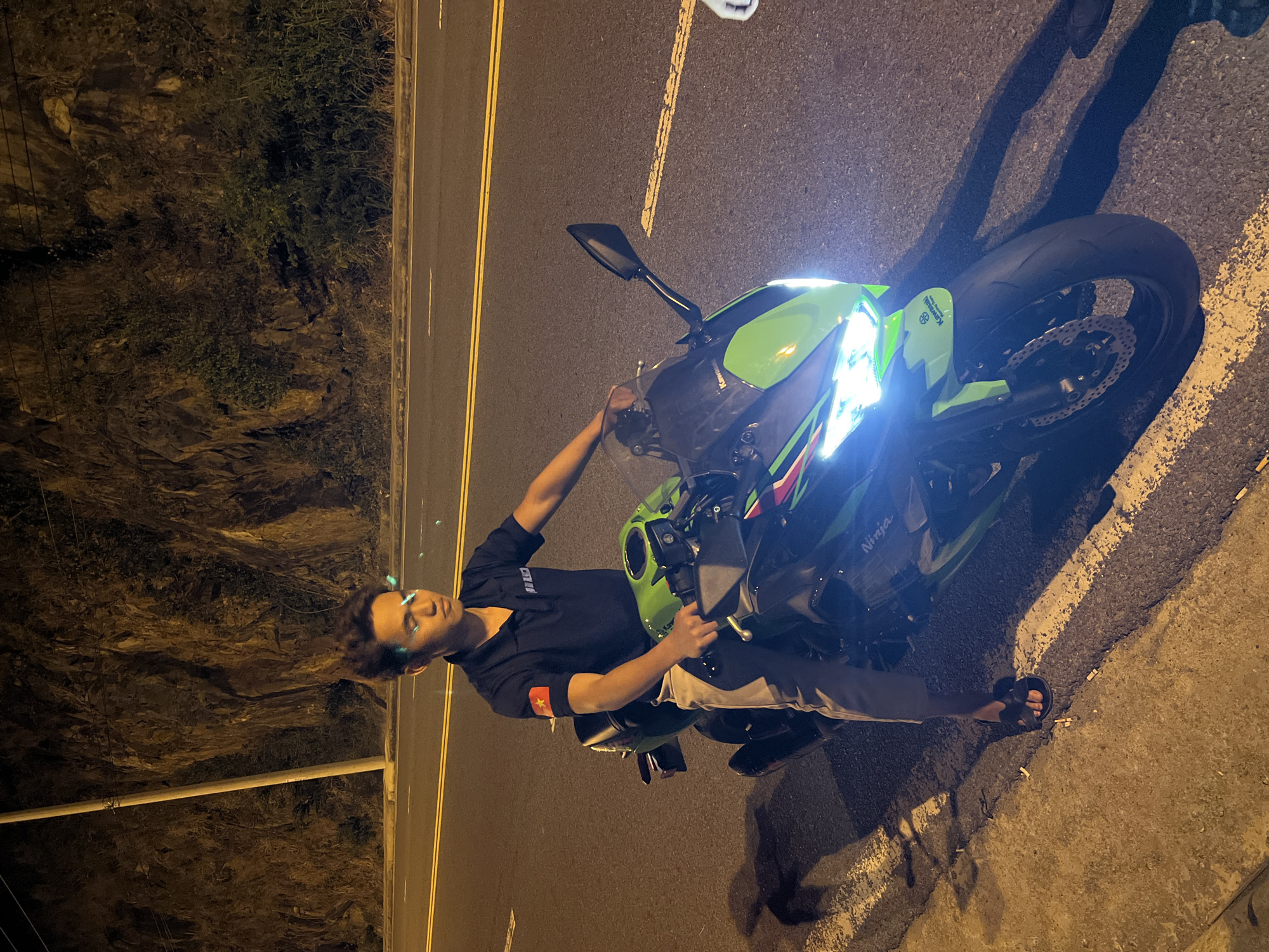 A dimly lit outdoor scene shows a person posing with a motorcycle at night, possibly on a roadside. The person is a young male dressed in a dark short-sleeved shirt with a patch on the upper arm, light-colored pants, and sandals. He has short dark hair and his facial features are illuminated by an indistinct light source. He is leaning slightly towards the front of a bright green and black Kawasaki Ninja motorcycle, its headlight prominently visible. The motorcycle is parked on a road shoulder next to a two-lane road with yellow center lines. Behind the person and the road is a rocky embankment covered with vegetation. The overall setting suggests an urban or semi-urban environment at night.