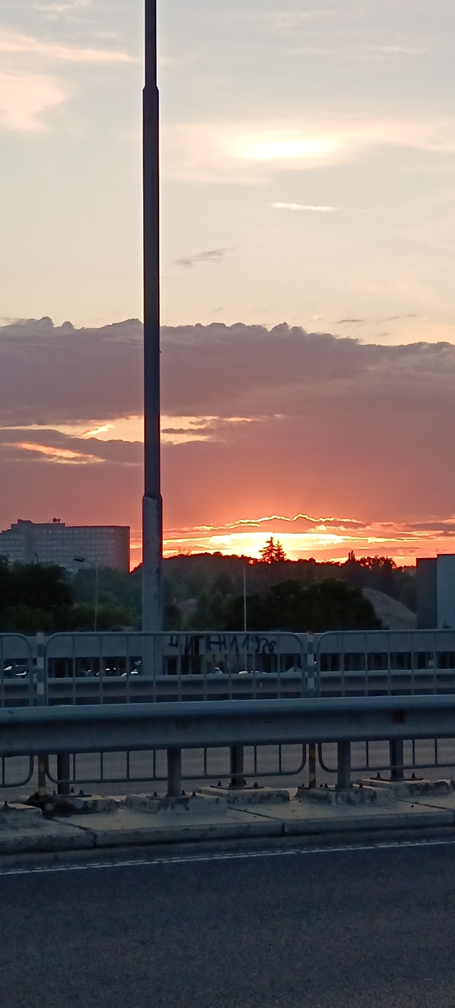 The image captures a vibrant sunset over an urban landscape, viewed from a road bridge in Plovdiv, Bulgaria. The sky is dominated by dramatic hues of orange and red near the horizon, gradually fading to lighter peach and soft grey higher up, with scattered clouds illuminated by the setting sun. A tall, dark pole, likely a street light or utility pole, stands vertically in the center, extending from the bottom to the top of the frame, silhouetted against the sky.
In the foreground, the asphalt road is visible at the very bottom, featuring a faint white dashed line. Above the road, a sturdy metal guardrail or barrier of the bridge spans horizontally across the lower third of the image. This barrier is composed of vertical posts supporting multiple horizontal rails and is anchored by concrete bases visible beneath it. On a concrete structure or wall behind the barrier, some visible graffiti includes Cyrillic text, reading "ЧИТИЛА 1976".
Beyond the barrier and stretching across the midground horizon, the silhouettes of trees and foliage line a low hill. To the left, a large, multi-story building with numerous windows is visible, also largely silhouetted against the bright sky. To the far right, the edge of another building is discernible. The setting suggests late evening, highlighting the beauty of the natural phenomenon amidst the constructed environment. No people are clearly visible, but the presence of the road and bridge implies an active urban environment.