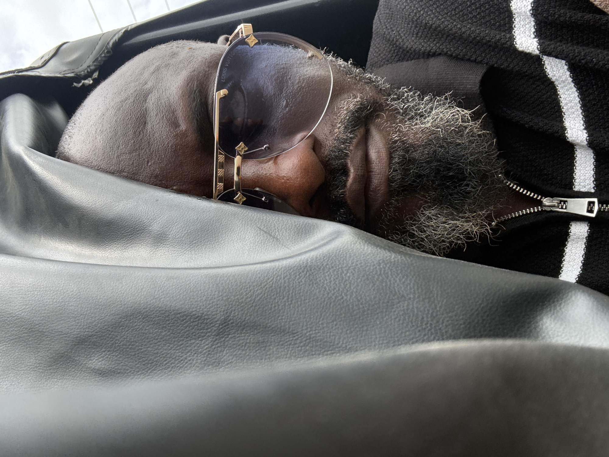 The image presents a close-up, slightly overhead view of an adult man in repose, likely sleeping or deeply resting. His dark-skinned, bald head is nestled against a black, textured material, possibly a leather or faux-leather seat or headrest. Large, round sunglasses with distinctive gold-colored frames rest on his face, obscuring his eyes. Faint text, "LOUIS VUITTON," is visible on the right lens of the sunglasses, along with characteristic brand motifs on the frame.
The man has a prominent, thick beard that is a mix of grey and white, curling around his jawline. He is wearing a black garment, possibly a jacket or sweater, featuring vertical white stripes and a silver zipper, visible on the right side of the frame. The background is indistinct, but a glimpse of a light, overcast sky can be seen in the upper left corner, suggesting daytime. The man appears peaceful and relaxed. The scene is set in Ado, Nigeria.