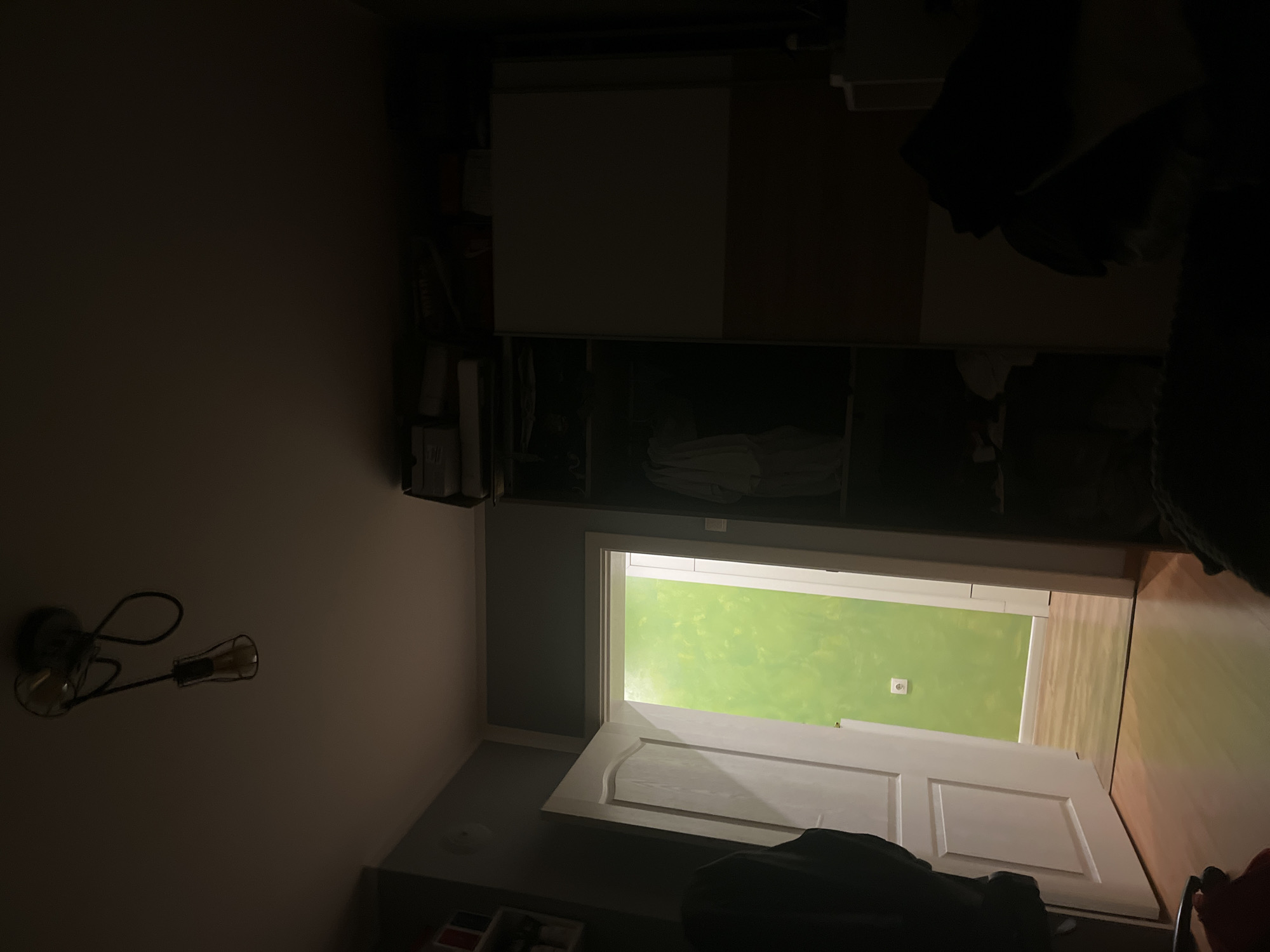 The image shows an interior view, likely in a residential setting, possibly located in Pokrovnik, Bulgaria (based on the prompt). The scene is dimly lit. A partially open white door leads to a room with bright green walls. A light fixture is visible on the left wall of the room. A wardrobe stands in the upper portion of the frame, appearing to have shelving and closed compartments. There are items stored on the shelves. In the foreground, obscured by shadows, is what appears to be a chair or other furniture. The overall composition suggests a bedroom or living space.