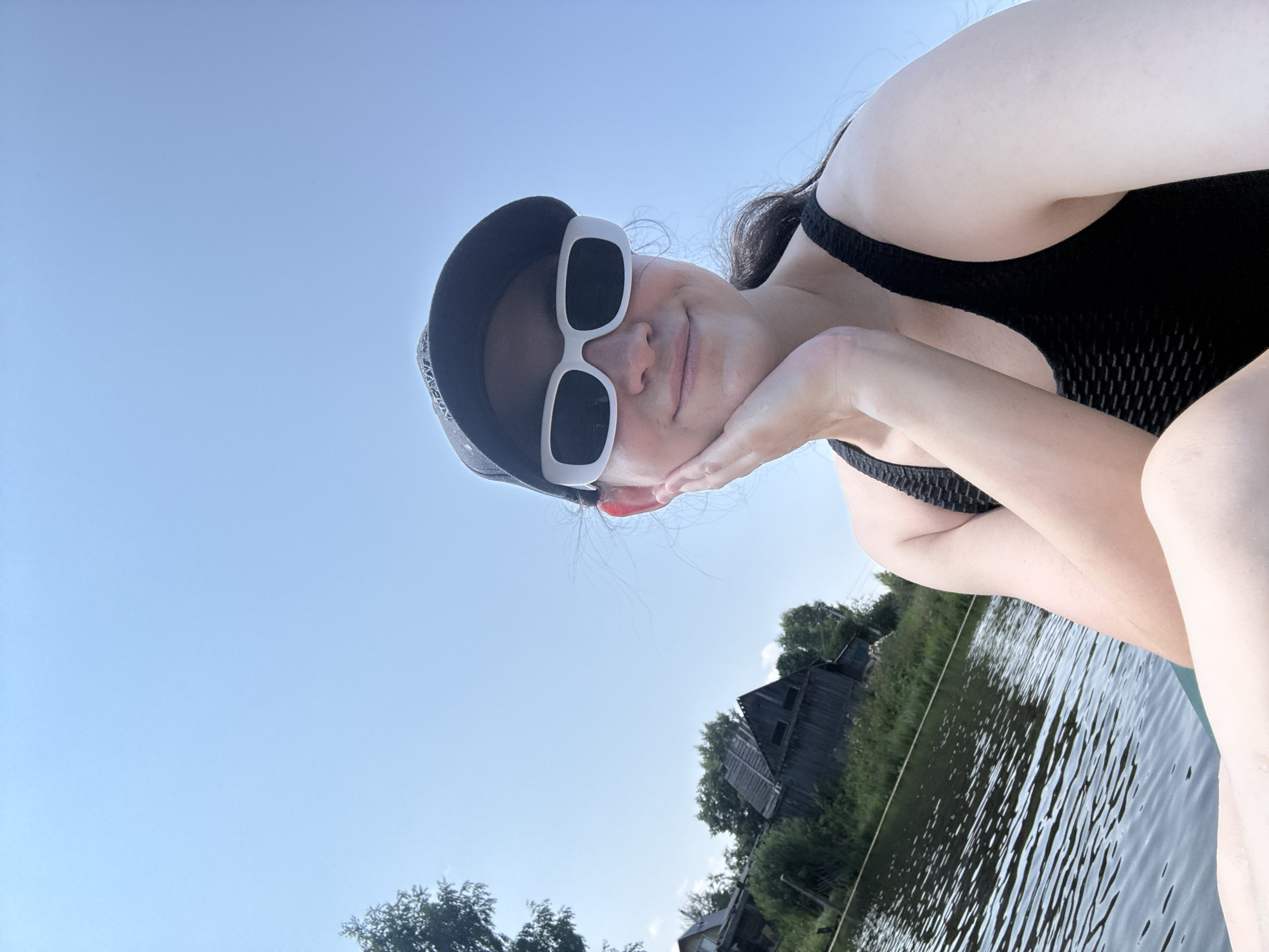 A young woman is taking a selfie from the perspective of a boat on a body of water, possibly a river or lake. She is wearing a black baseball cap, white rectangular sunglasses, and a black tank top. Her right hand is resting on her chin, and she has a slight smile. The background shows a clear blue sky, a line of trees and vegetation along the bank, and several rustic wooden buildings. The water in the foreground is rippled, indicating a gentle breeze. The overall scene suggests an outdoor recreational activity on a sunny day, possibly in a rural or semi-rural area of Zasechnyi, Russia.
