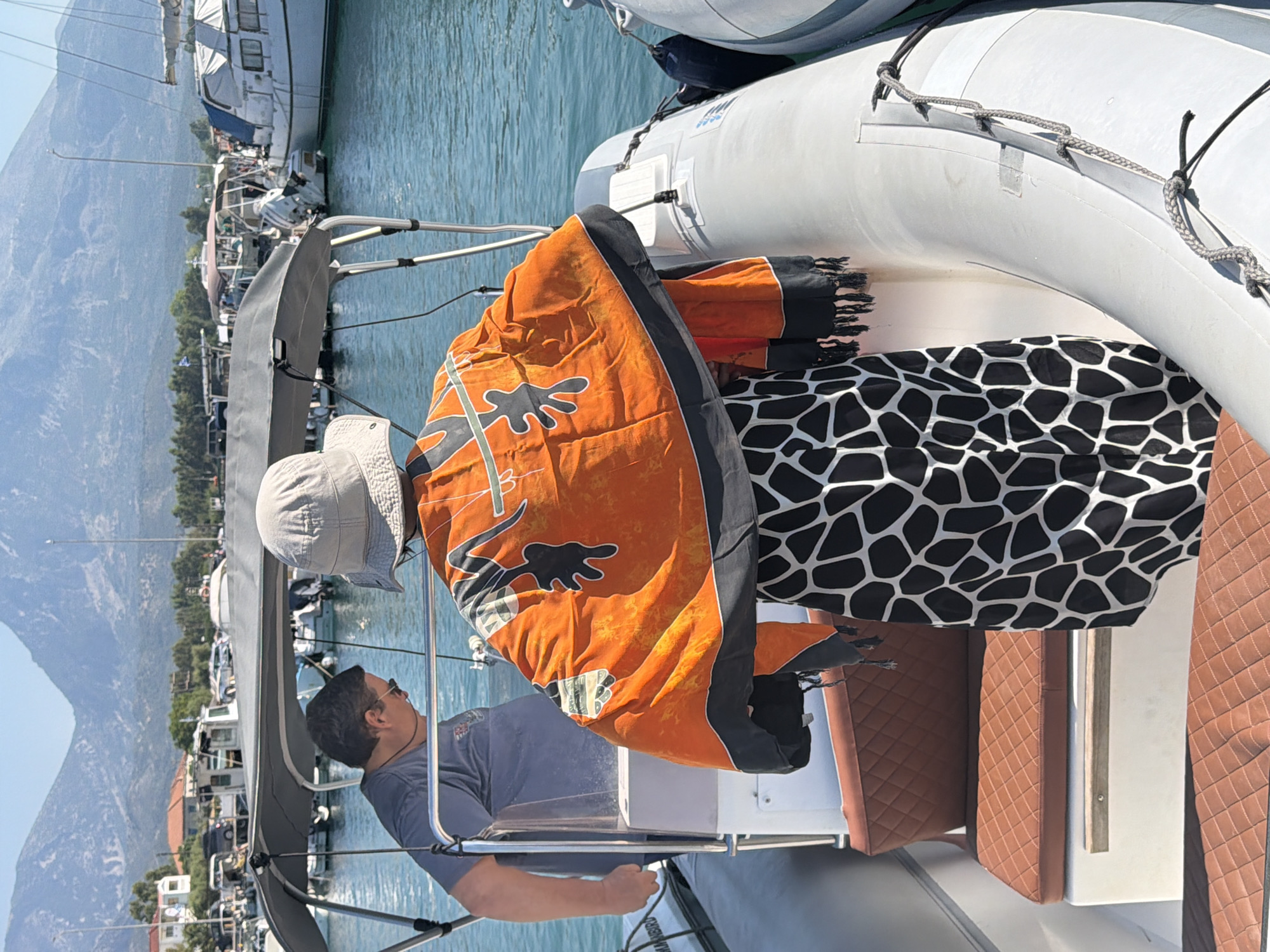 In the Municipal Unit of Alyzia, Greece, two individuals are on a boat docked in a harbor. A man wearing a grey t-shirt and sunglasses stands on the left, facing away from the camera. In front of him, another person, whose face is not visible, is turned away from the camera, wearing a light-colored hat and an orange garment with black animal print patterns draped over their shoulders. This garment has a black fringe at the bottom. The boat has brown quilted seating visible. In the background, several other boats and a waterfront town with buildings and mountains can be seen under a clear blue sky.
