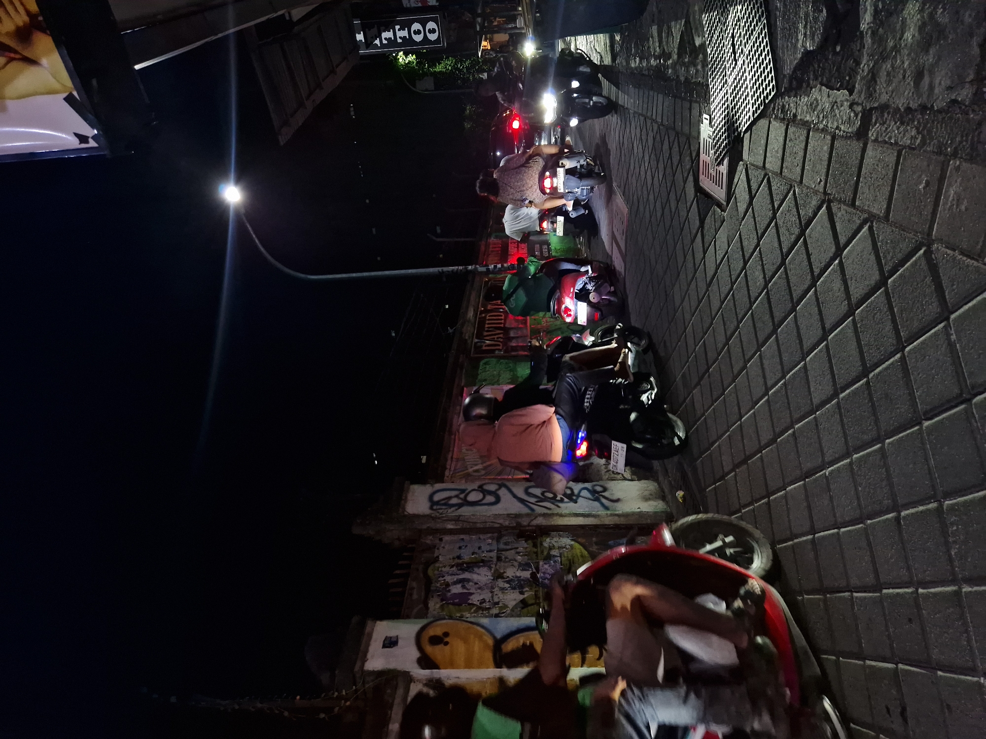 At night in Kuta, Indonesia, a line of motorbikes is stopped on a narrow street next to a graffiti-covered wall. The street is paved with textured grey bricks. Above the motorbikes, a street lamp illuminates the scene. A few pedestrians are also visible. The storefronts are to the right, with a tattoo shop sign visible.