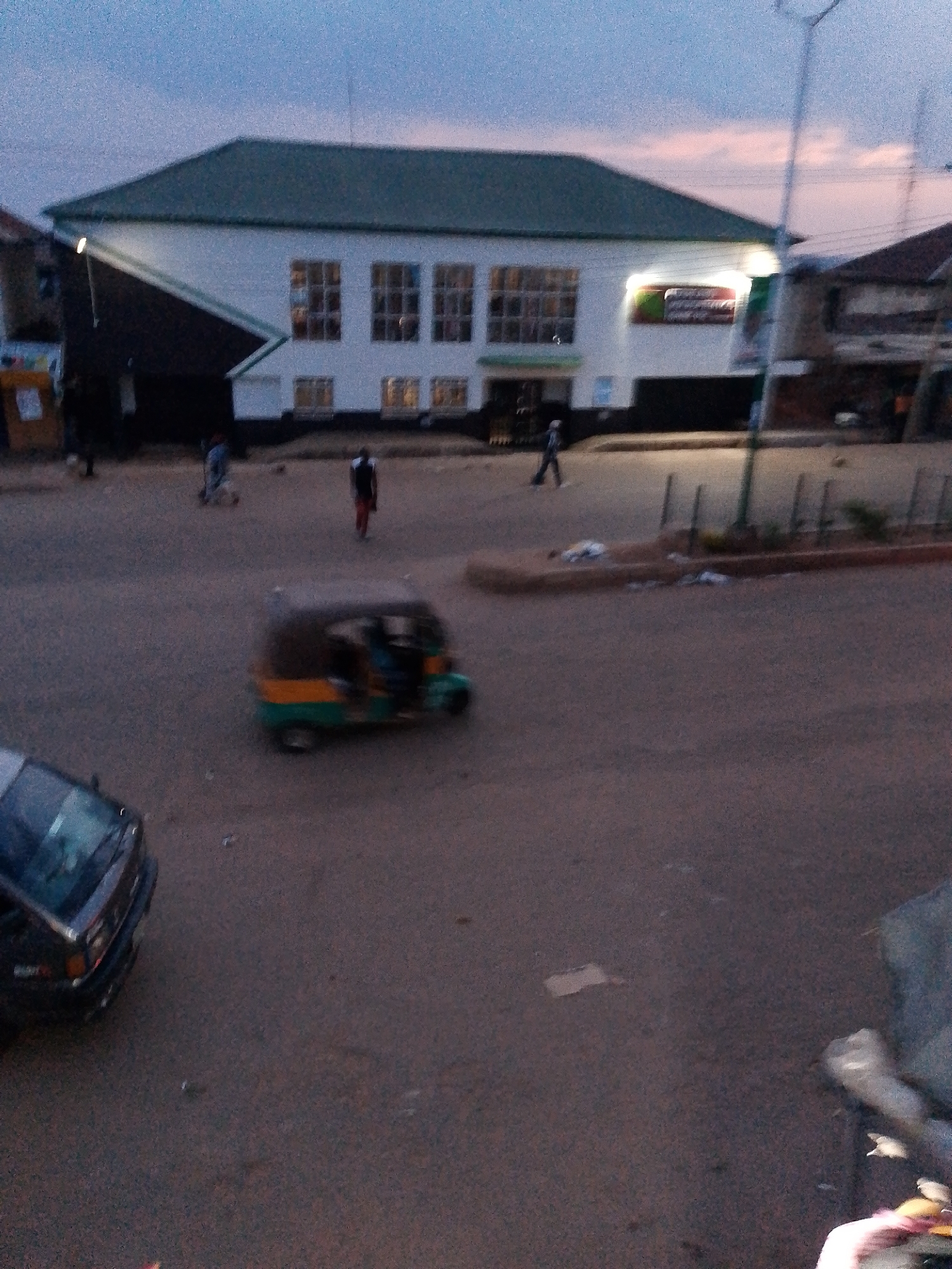 The image depicts a street scene at dusk in Bukuru, Nigeria. In the background, a white building with a green roof is visible. The building has several large windows and a prominent sign with red and green lettering. To the left of the building, another structure is partially visible. In the mid-ground, a three-wheeled auto-rickshaw, commonly known as a "Keke Napep," is in motion. Its body is painted green and yellow. A dark-colored car is parked on the left side of the foreground. Several individuals are present in the scene: one person is walking in the mid-ground, another is walking on the right side, and two people are visible inside the Keke Napep. The ground is paved, and there is some debris scattered on the road. The sky is a dusky blue with hints of pink. The overall lighting suggests it is either early morning or late evening.