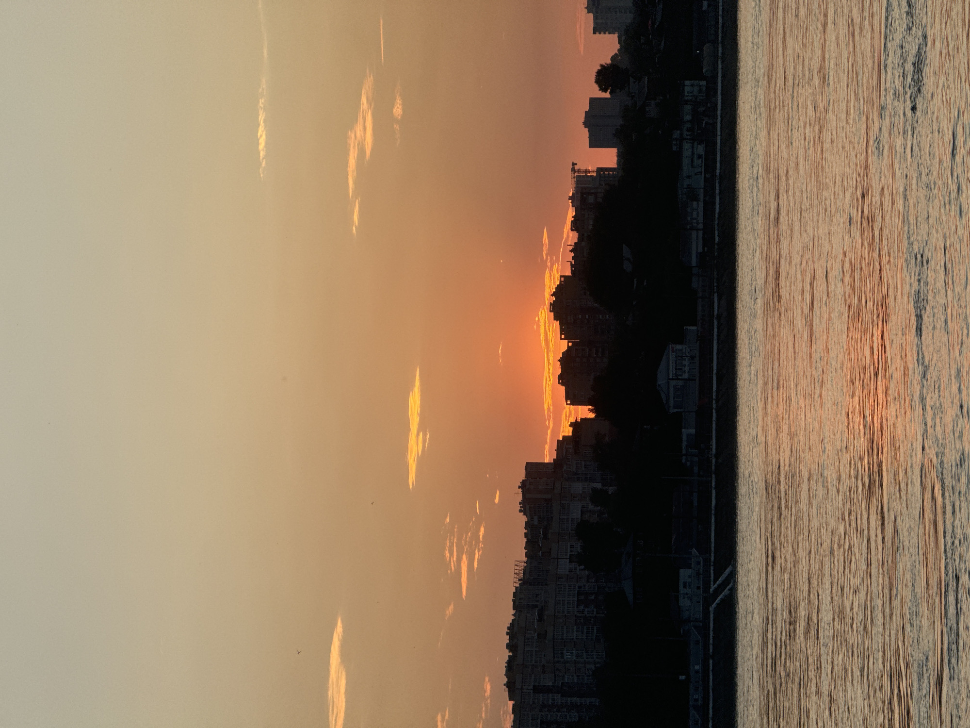 This is a vertical, medium-shot photograph of a sunset over the Angara River in Irkutsk, Russia. The sky is a gradient of warm orange and yellow hues, with scattered, wispy clouds. The setting sun is visible as a bright orange orb peeking through a gap in the clouds, casting a warm glow. In the mid-ground, the silhouette of a city skyline is visible against the bright sky, characterized by several apartment buildings of varying heights and architectural styles. Trees are silhouetted against the buildings, obscuring the lower portions of the structures. A construction crane is visible on the right side of the skyline. The foreground is dominated by the Angara River, its surface rippled by gentle waves that reflect the warm colors of the sunset, creating a shimmering effect. The riverbank is visible in the lower mid-ground, with a dark, silhouetted embankment and some indistinct structures along the edge. The overall impression is one of a tranquil evening scene in an urban environment.