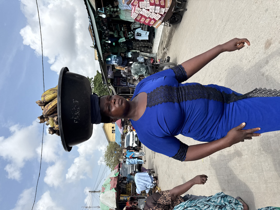 The image displays a bustling outdoor market street in Lagos, Nigeria, under a partially cloudy sky. The primary subject is an adult woman with dark skin, centrally positioned, walking towards the viewer's right. She wears a royal blue, knee-length dress featuring black lace trim along the front and sleeves. On her head, she carries a dark, weathered plastic basin filled with numerous plantains or large green bananas, some exhibiting brown discoloration, balanced on a folded blue fabric.
In the mid-ground, to the left of the central figure, a person in a light blue garment bends over near a pushcart. Additional pedestrians and parked vehicles, including a white car, are visible further down the street, beneath market stalls with green and white striped awnings. A yellow building is discernible in the distant left.
To the right, a series of open-front stalls line the street. One stall prominently displays clothing, including t-shirts and trousers, on several mannequins. A person on a motorcycle is visible in front of these stalls. Further right, a large handcart, equipped with two wheels, is heavily loaded with multiple stacks of rectangular red and white boxes. Utility lines traverse the upper portion of the frame. The ground is a light-colored, paved surface.