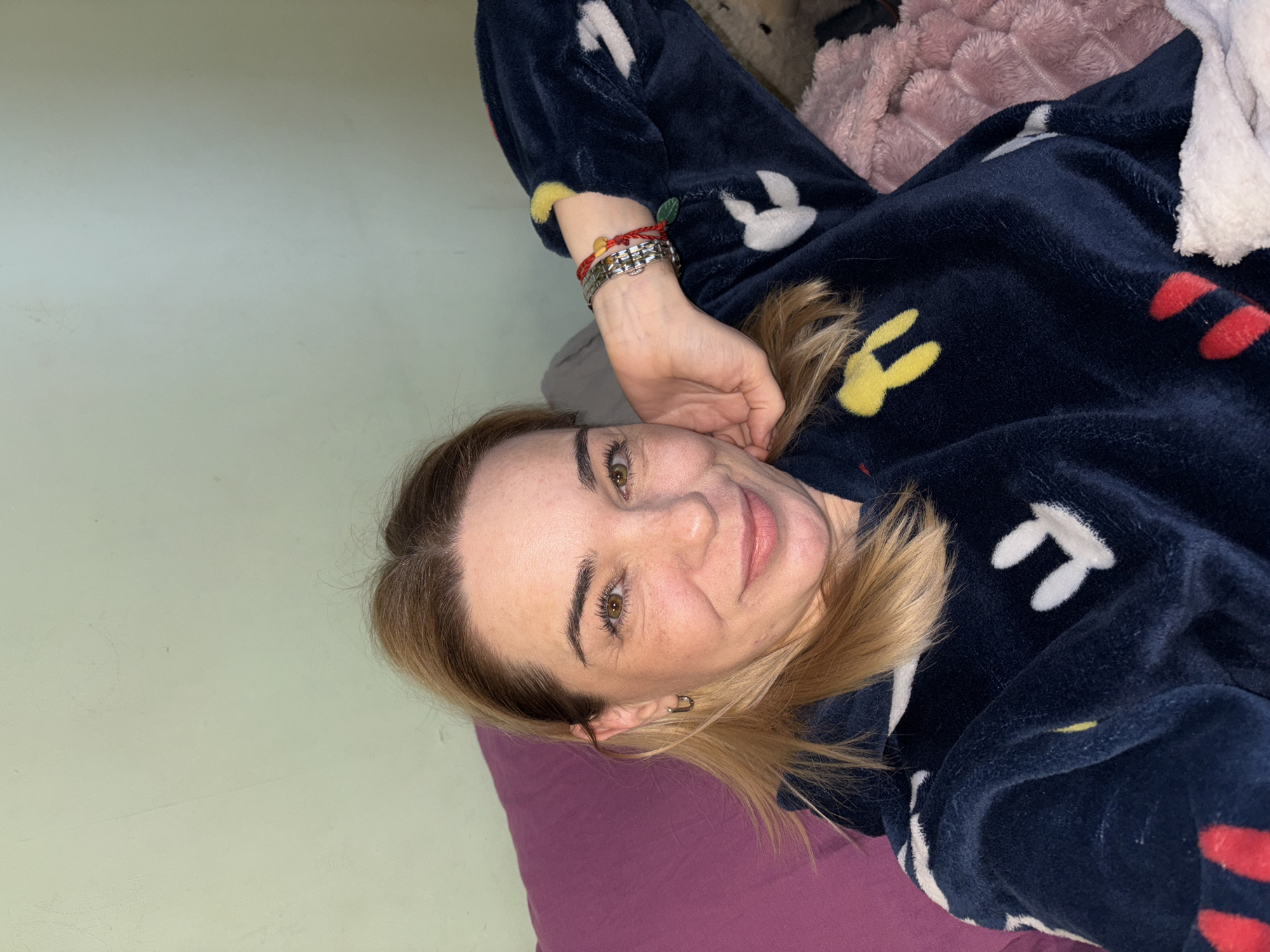 The image shows a woman lying on a bed. She is wearing a dark blue pajama top with cartoon character designs. She has fair skin, brown hair with blonde highlights, and green eyes. Her left hand is raised near her face, and she is wearing bracelets on her left wrist. The bed has a purple pillow and a pink and white blanket. The background includes a light green wall. The location is unknown. The city is Unknown, Russia.